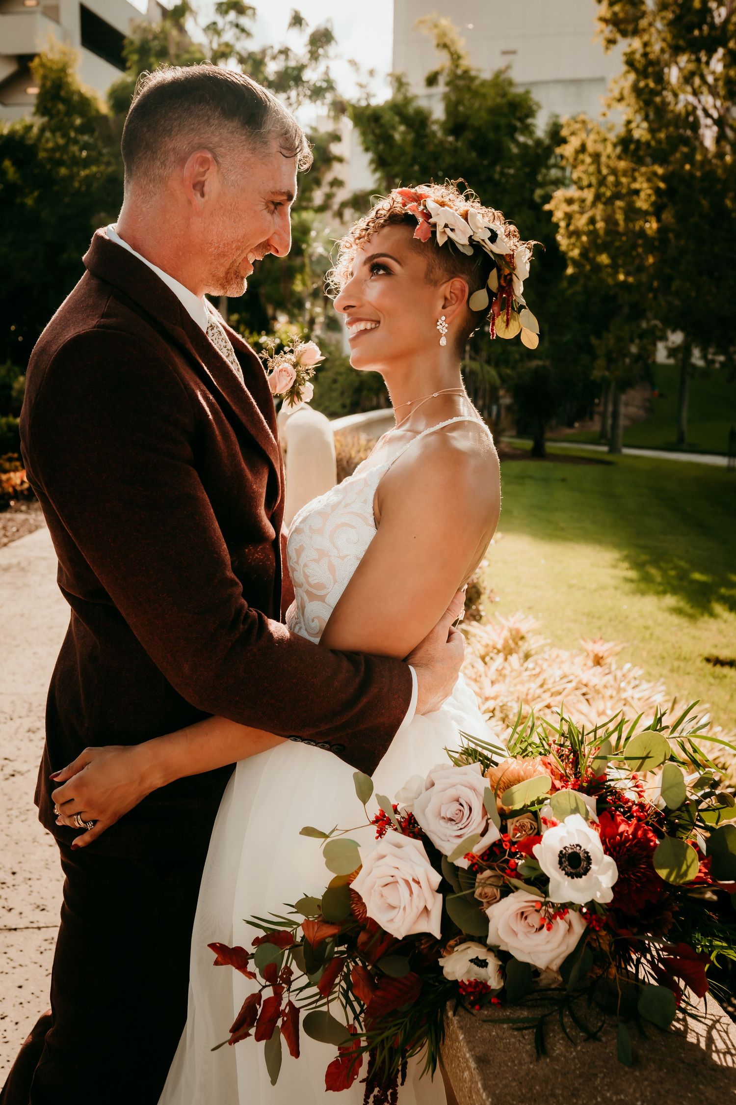 Bride and groom embrace outdoors, she wears a floral crown and holds a bouquet of red, white, and pink flowers.