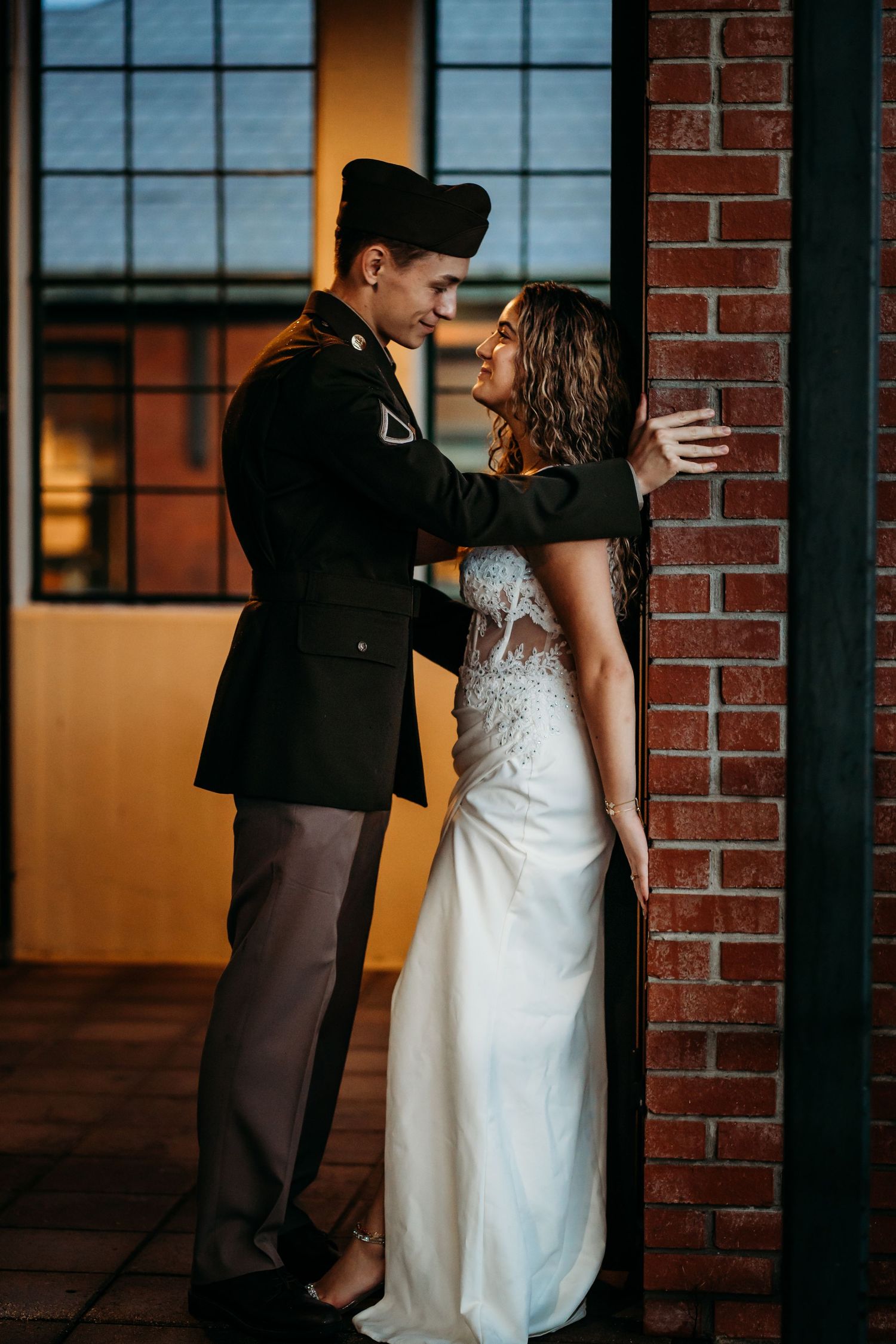 Military groom in uniform embracing bride in white wedding dress near brick wall in romantic indoor setting.