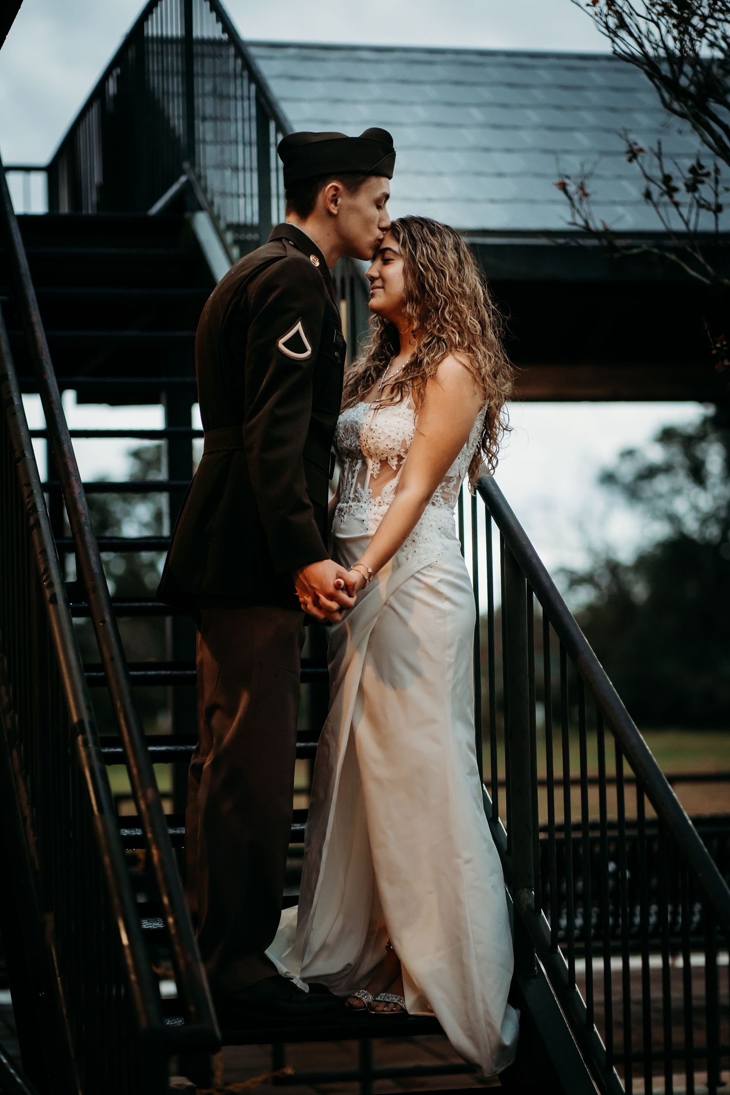 Military groom in uniform and bride in white dress holding hands on outdoor metal staircase.