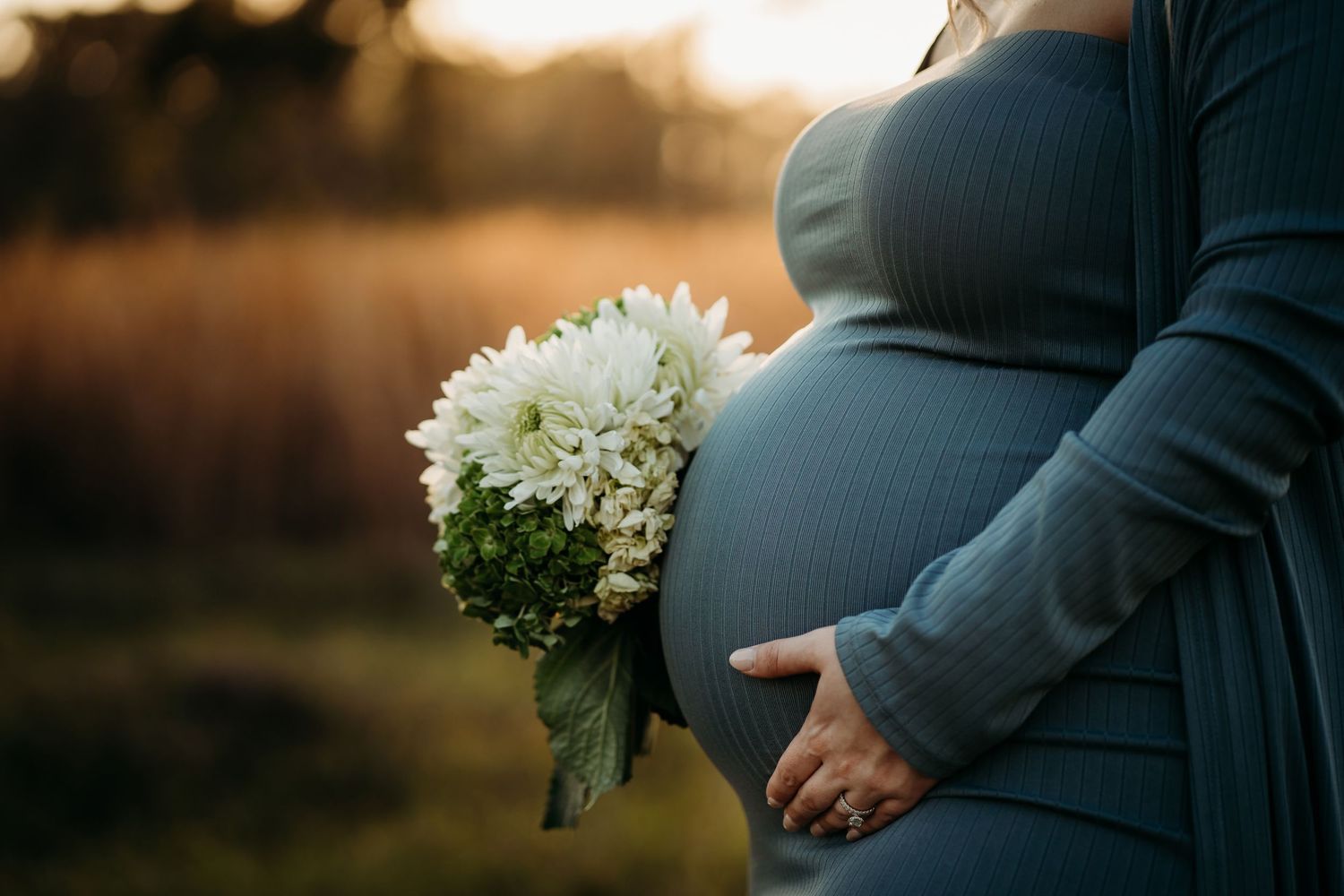 Close-up of pregnant belly in blue dress with white flower bouquet at sunset outdoors.
