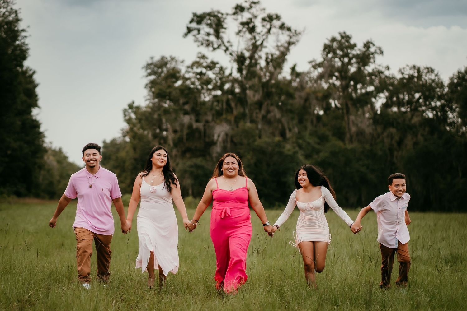 Five people in pastel outfits hold hands running through a green field with trees in the background.