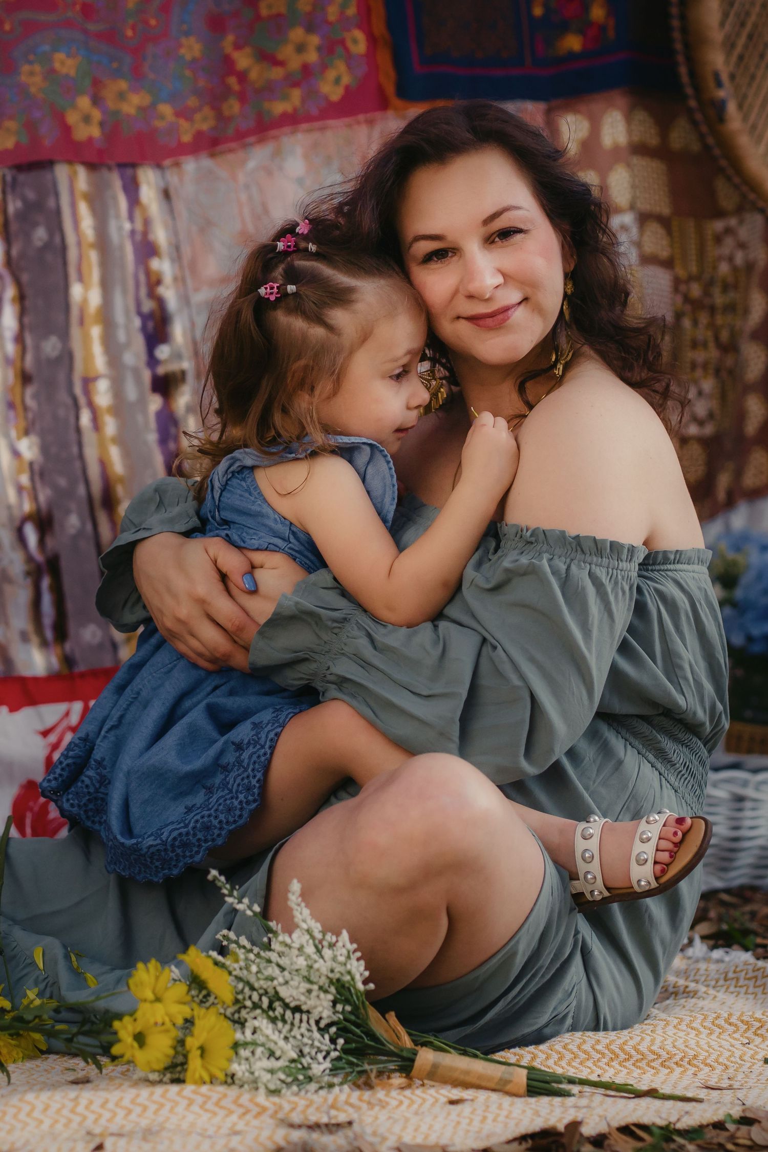 A woman in a gray off-shoulder dress holds a little girl in a blue dress, sitting together in a boho-styled setting.