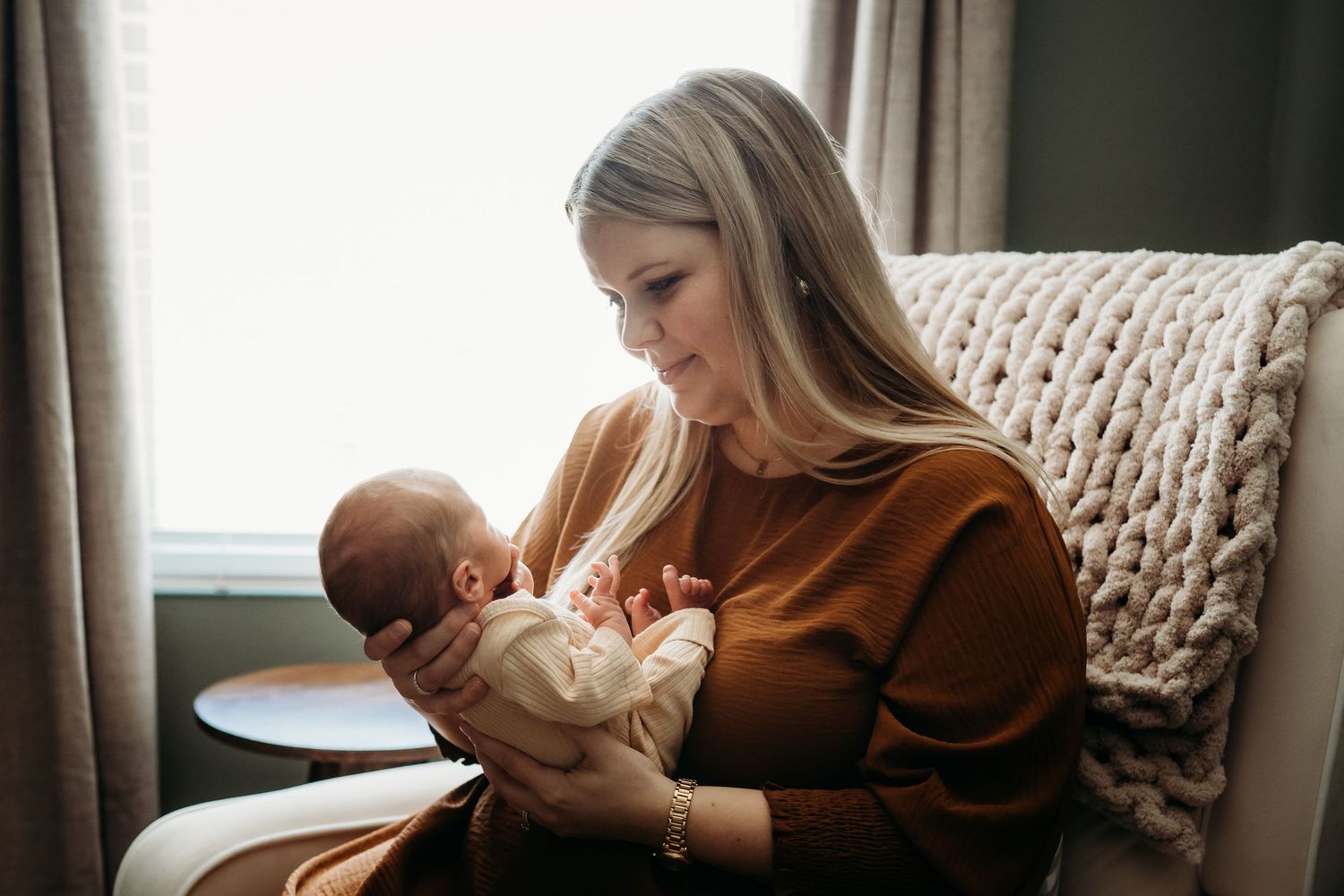 New mother in rust dress sitting in armchair, lovingly holding her newborn baby indoors.
