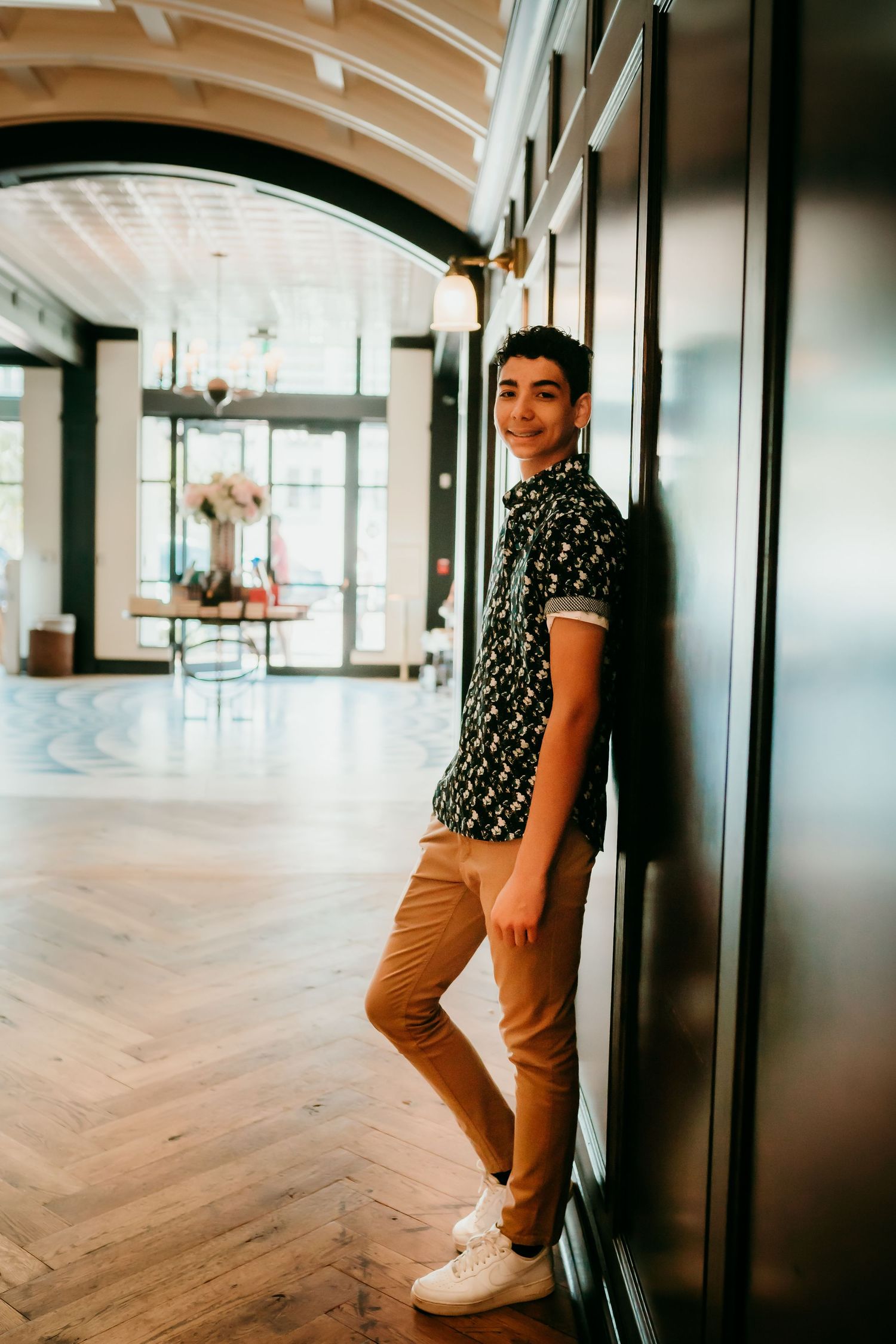 Young man in floral shirt and tan pants leaning against dark wall in elegant hotel lobby.