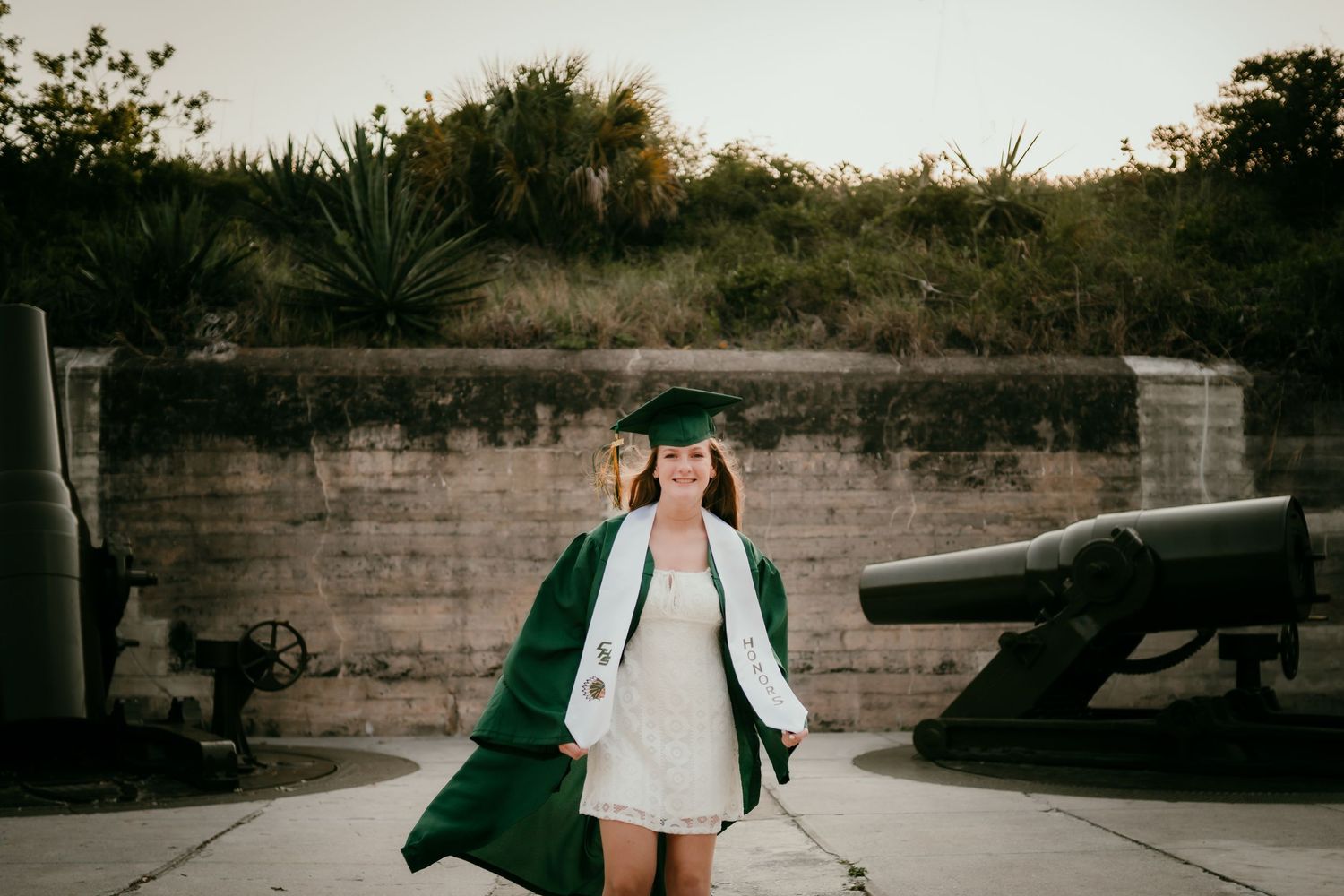 A smiling graduate in green cap and gown poses outdoors near historic cannons and stone walls.