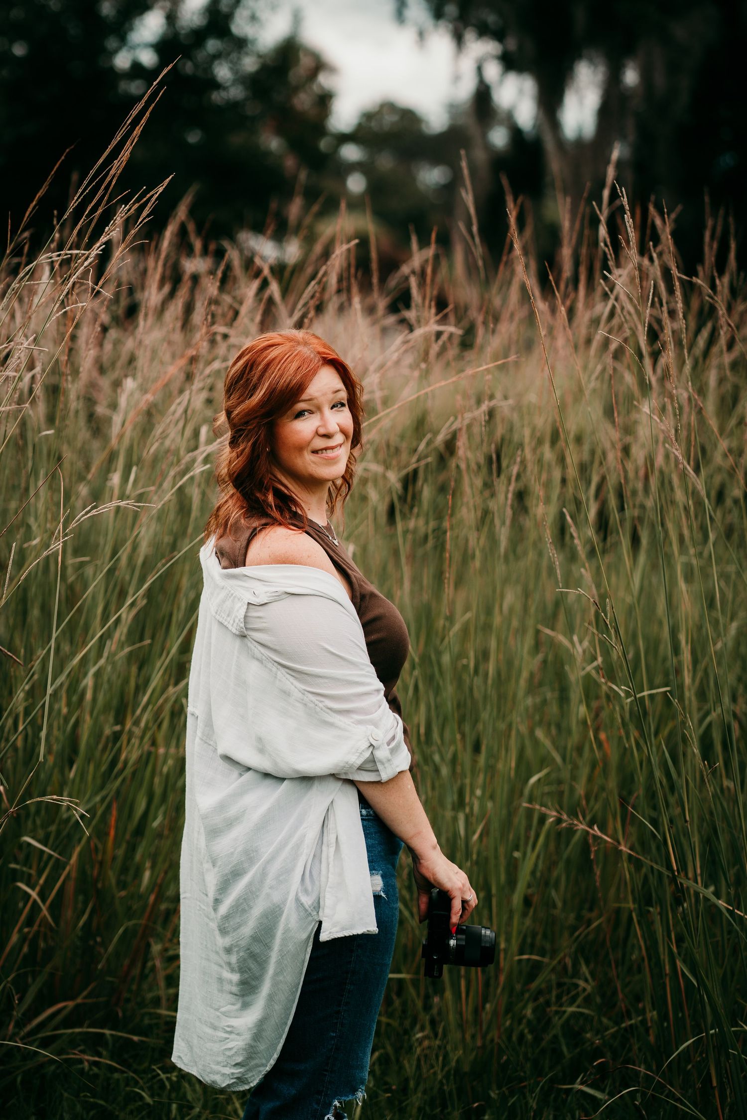 A red-haired woman in a white shirt holds a camera while standing in tall grass fields, smiling over her shoulder.