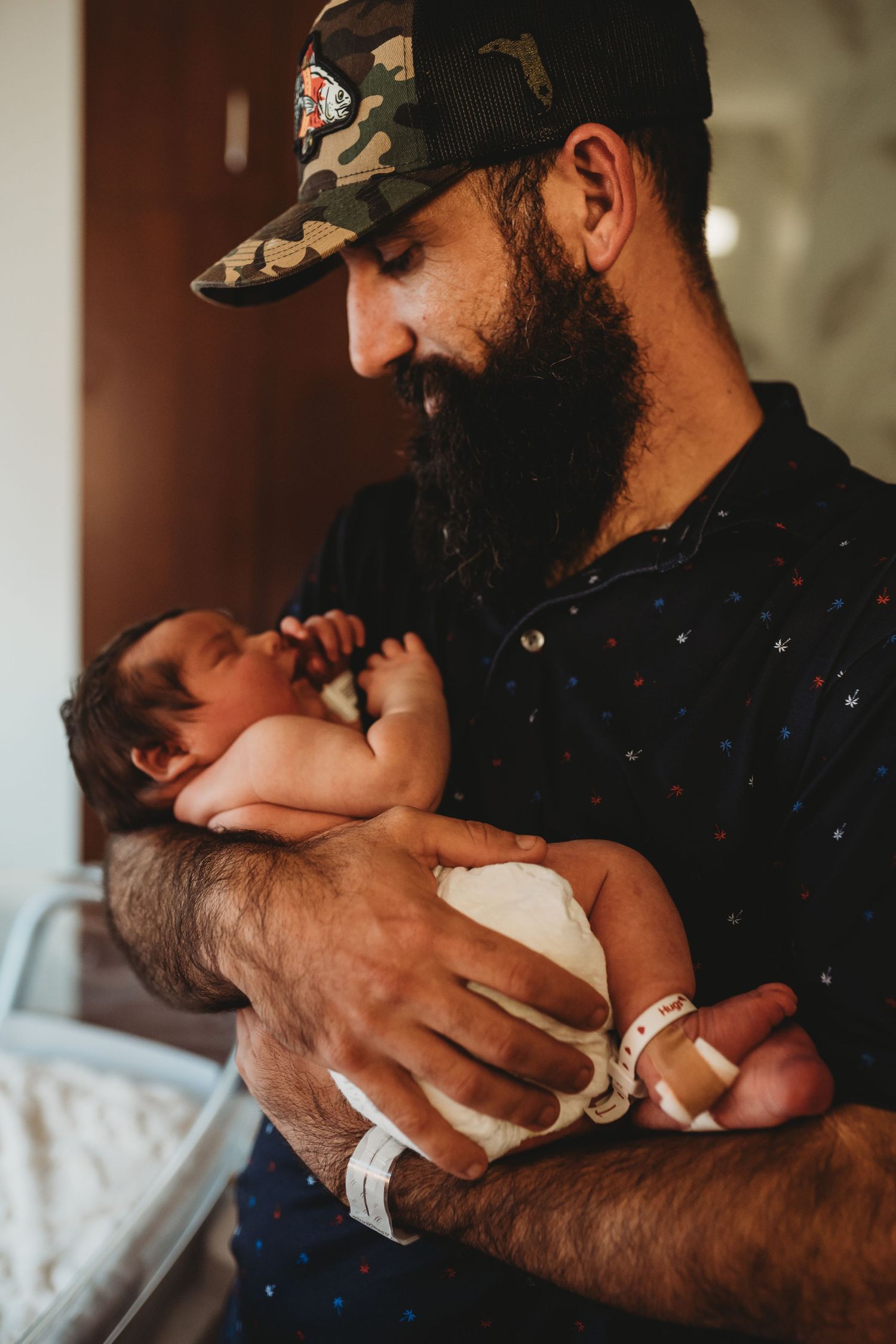 Bearded father in camo hat tenderly holding newborn baby in hospital room.