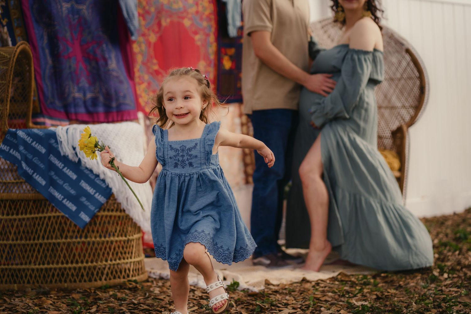 A joyful toddler girl in a blue denim dress runs outdoors during a boho family maternity photo session.