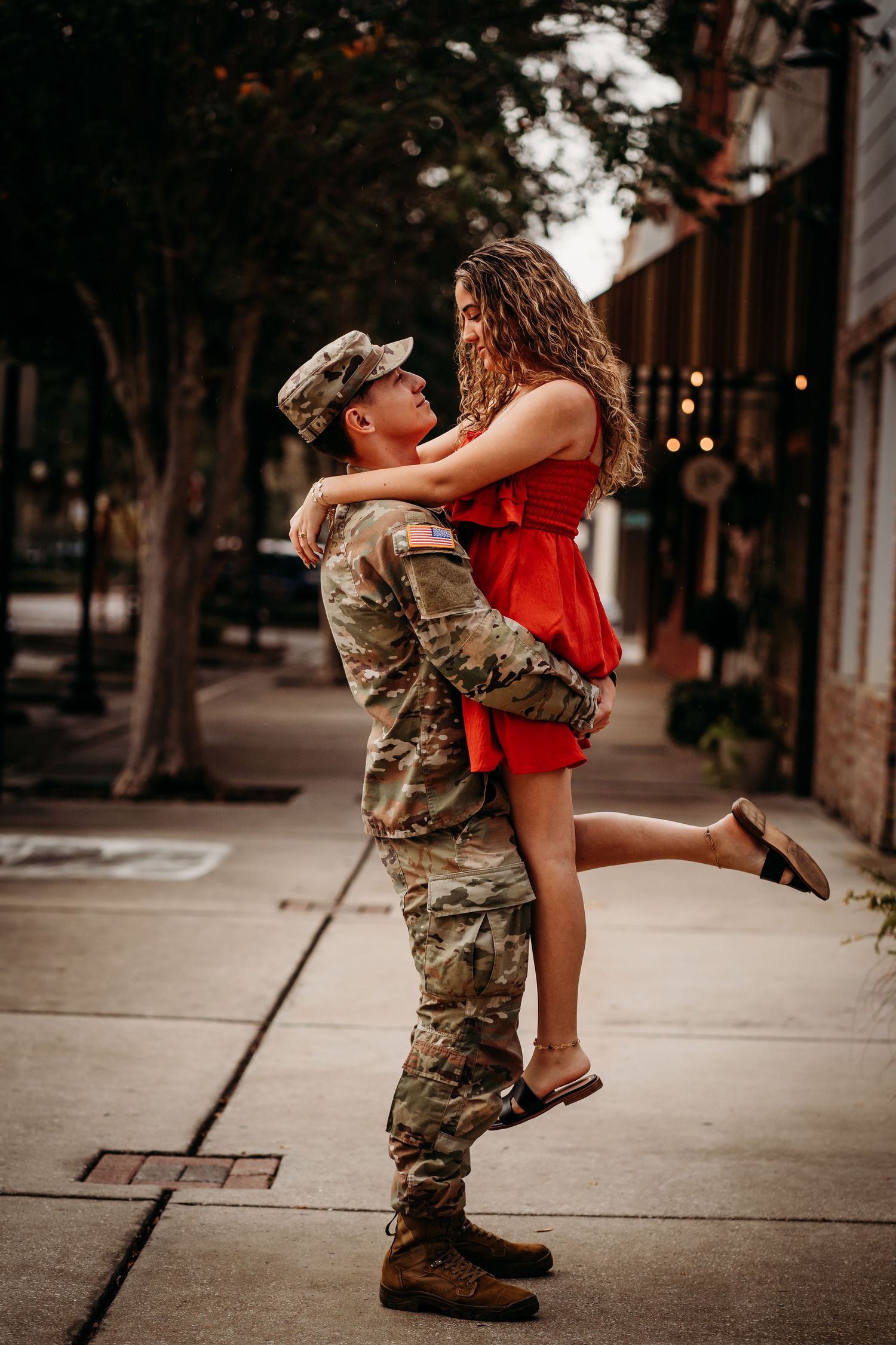 US Army soldier in camouflage uniform lifting woman in red dress on a city sidewalk.