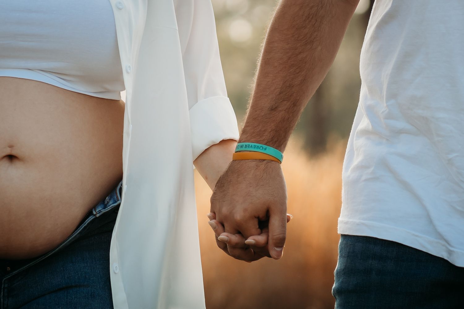 Couple holding hands at sunset, man wearing colorful wristband, woman showing baby bump.