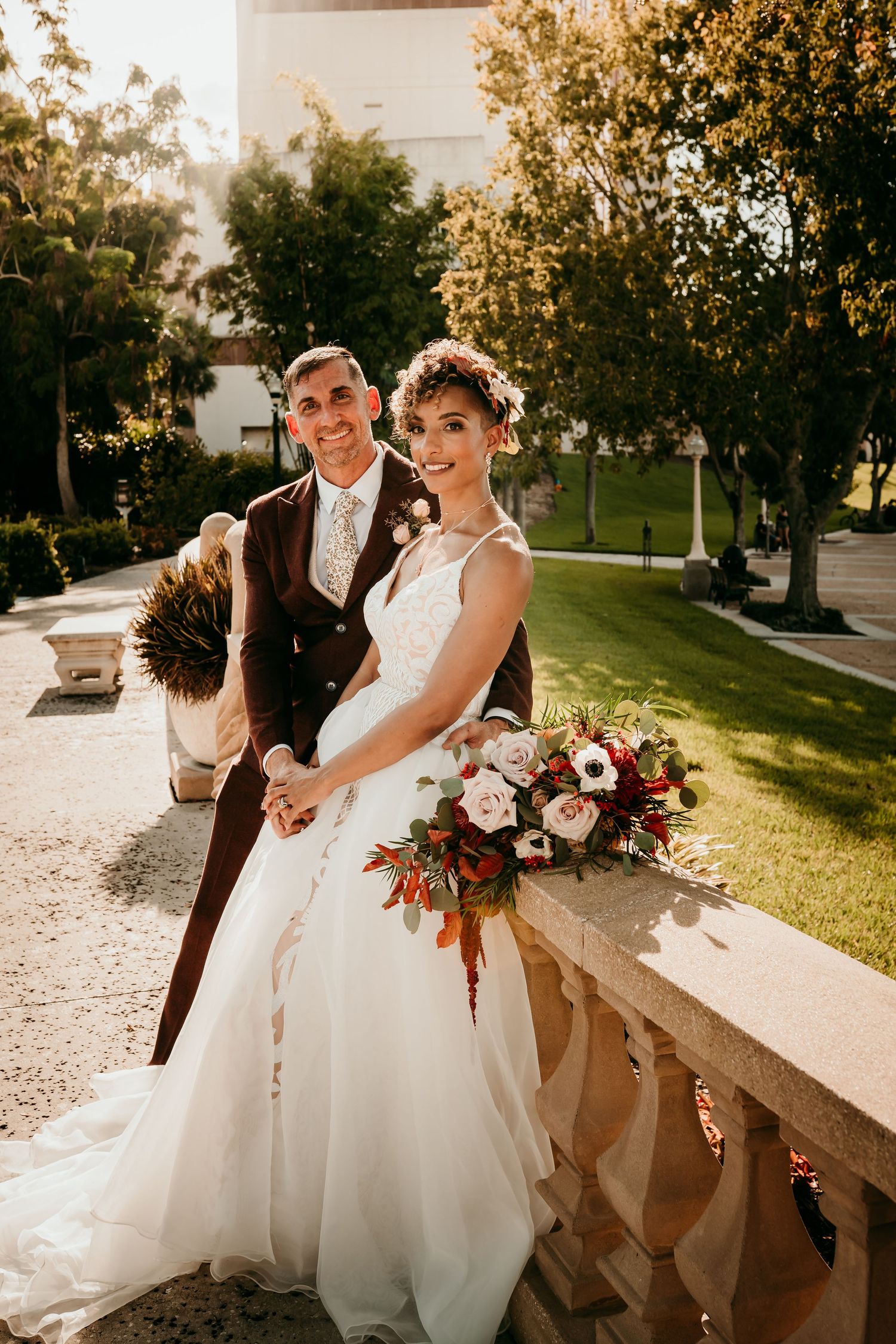 Bride and groom pose outdoors holding a red and white bouquet, wearing wedding attire in a sunlit garden setting.