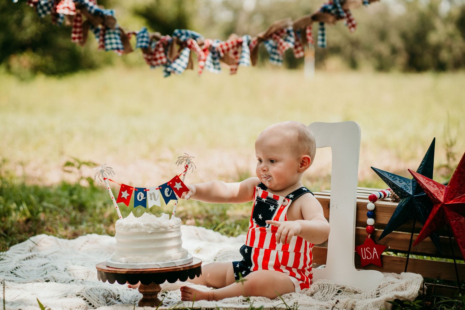 Baby in patriotic outfit celebrates first birthday with white cake and American-themed decorations outdoors.