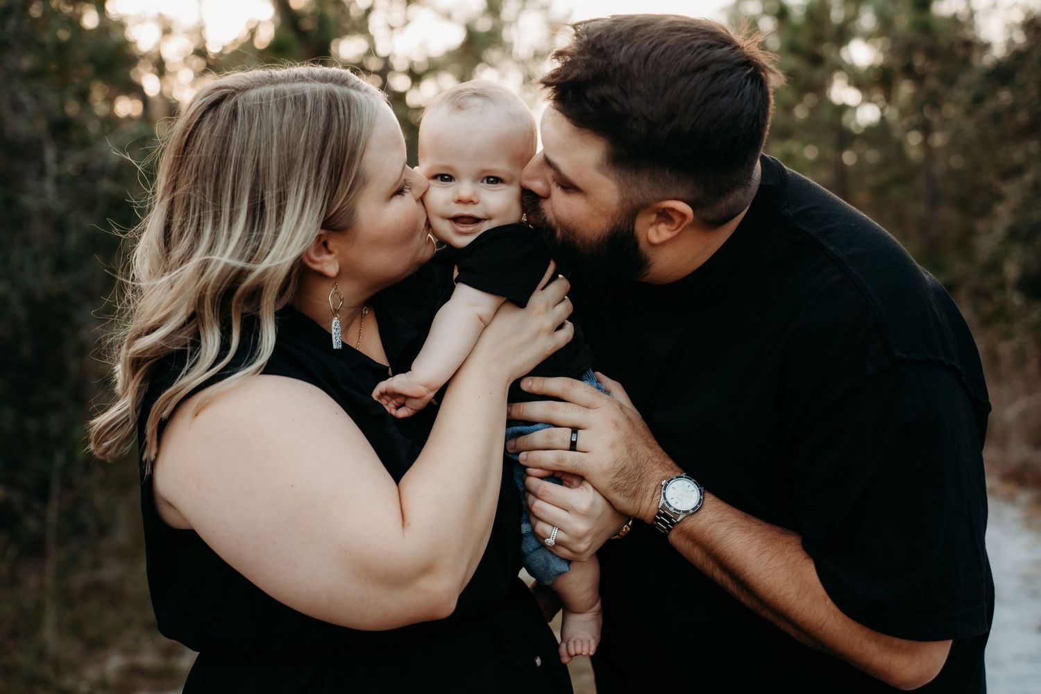 Happy parents kissing their smiling baby on the cheeks in a warm outdoor portrait.