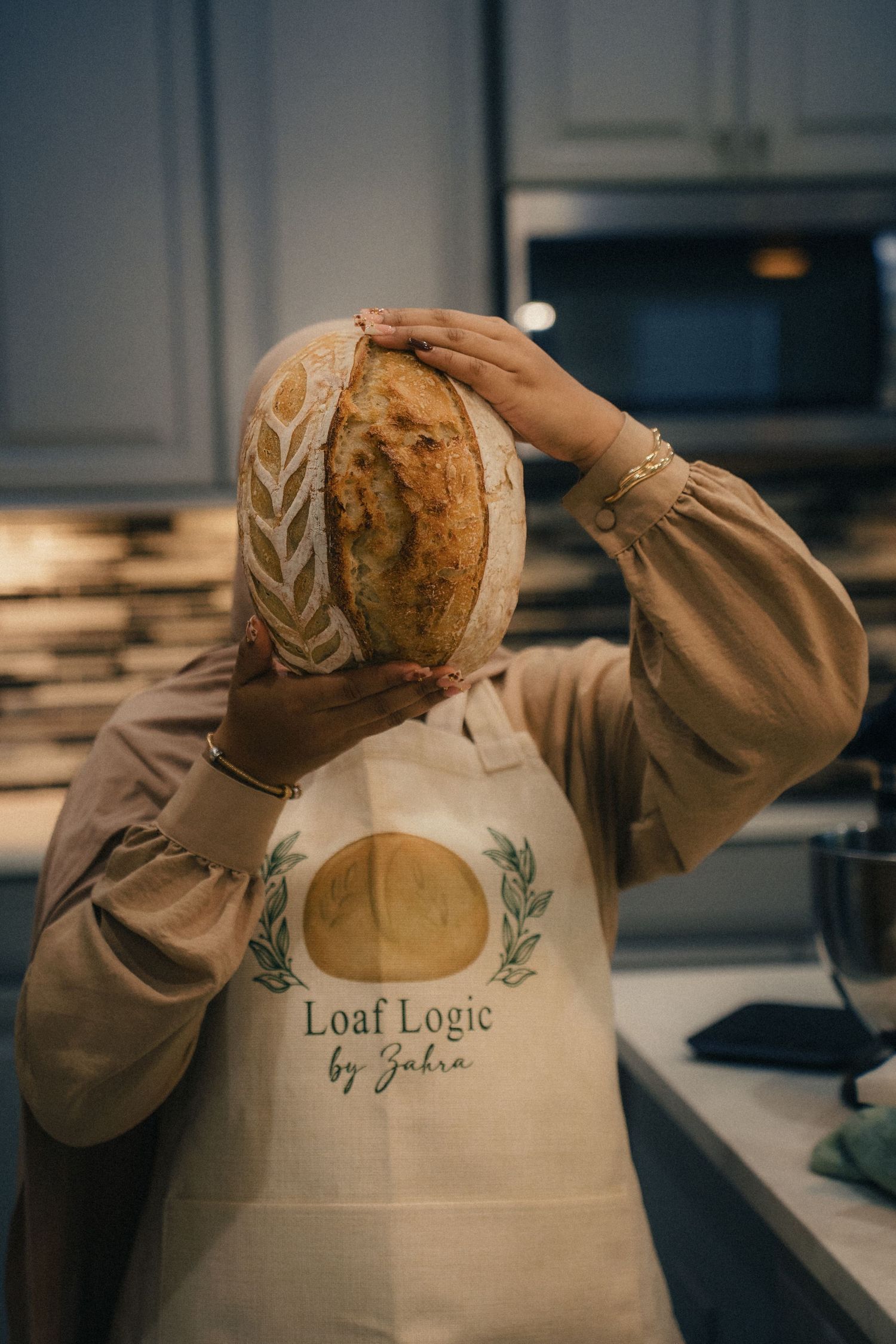 Baker holding sourdough bread loaf wearing a Loaf Logic by Zahra apron in a modern kitchen.