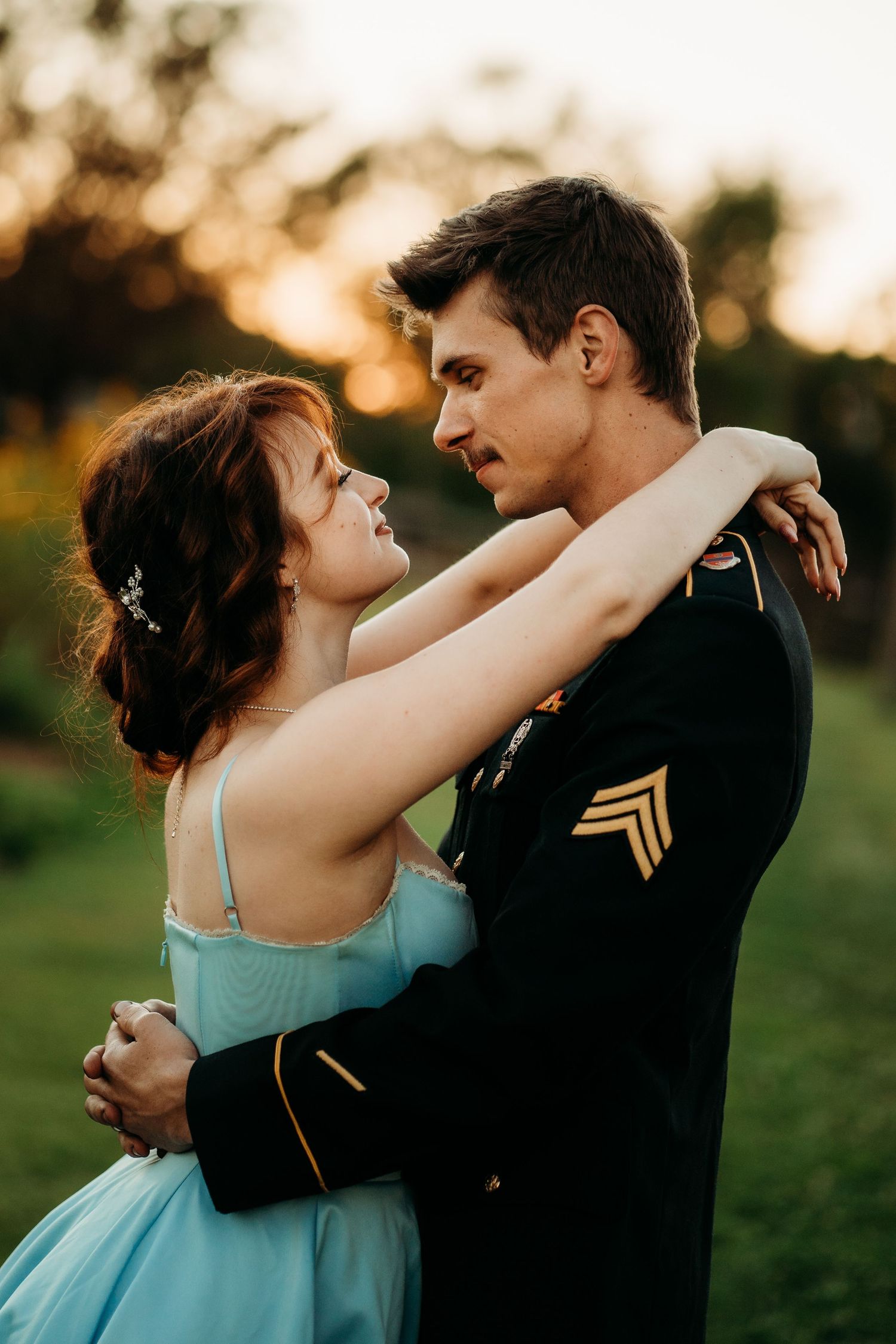 Couple slow dancing outdoors at sunset, woman in blue gown embraces soldier in Army dress uniform.