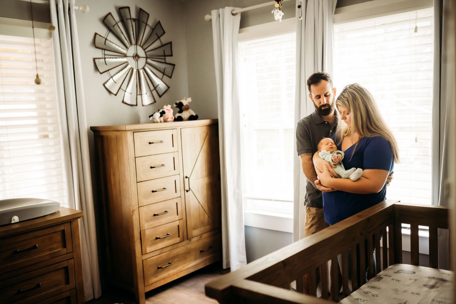 New parents admiring their sleeping newborn baby near wooden dresser in decorated nursery.