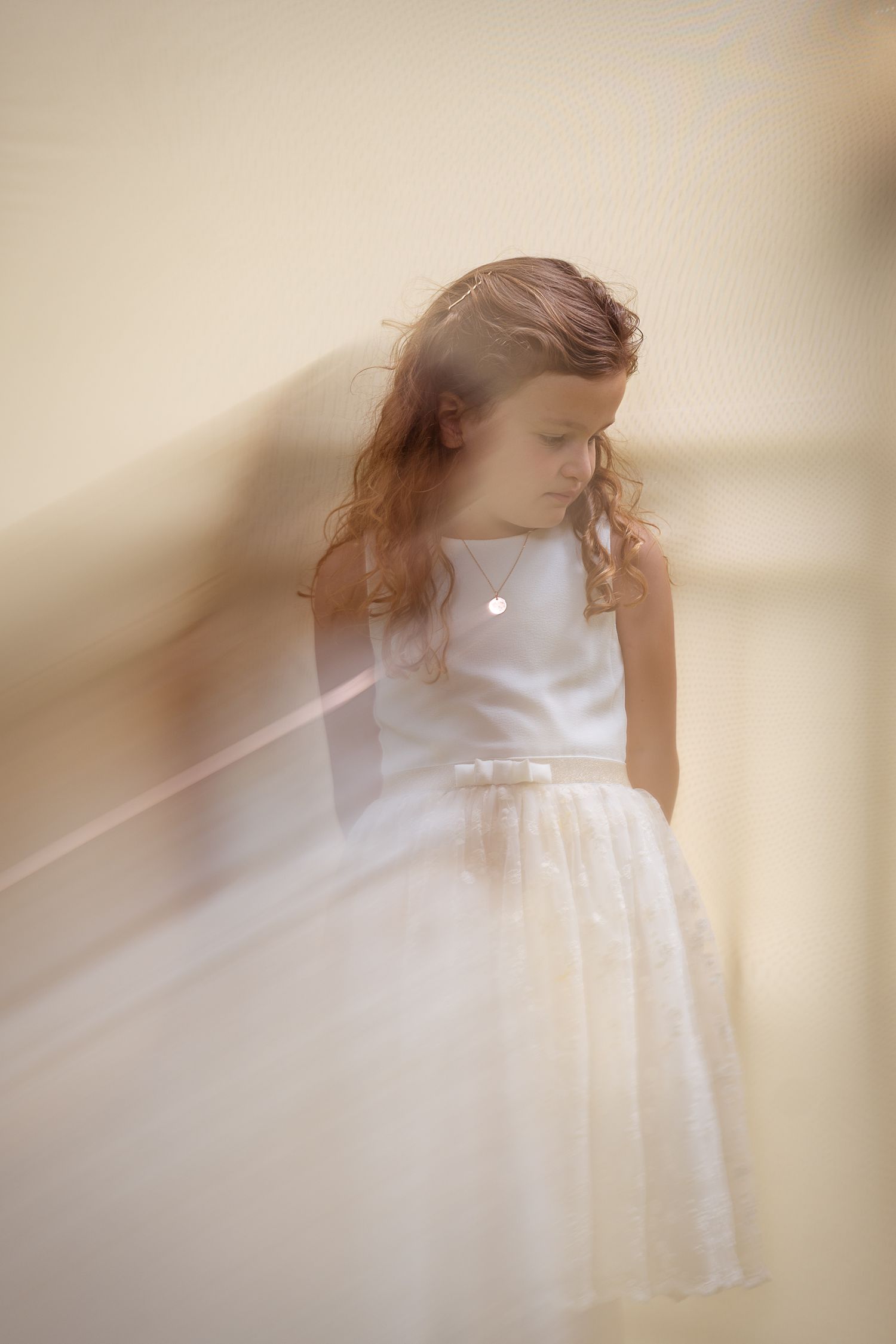 Young girl in white communion dress standing against a wall in soft dreamy light.