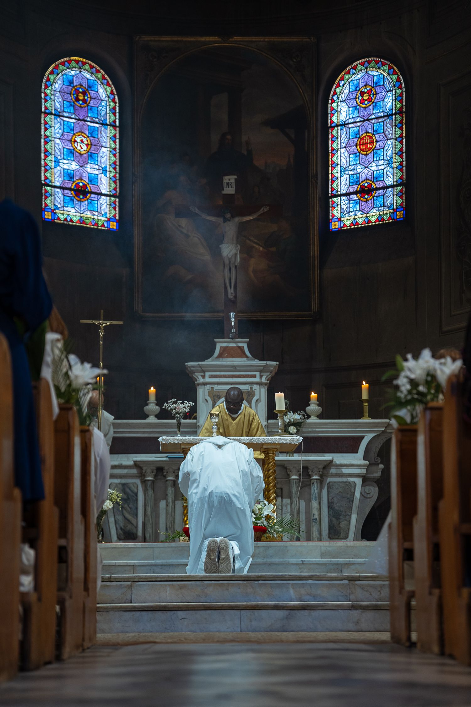 Catholic priest kneeling at ornate church altar with stained glass windows and crucifix during mass.