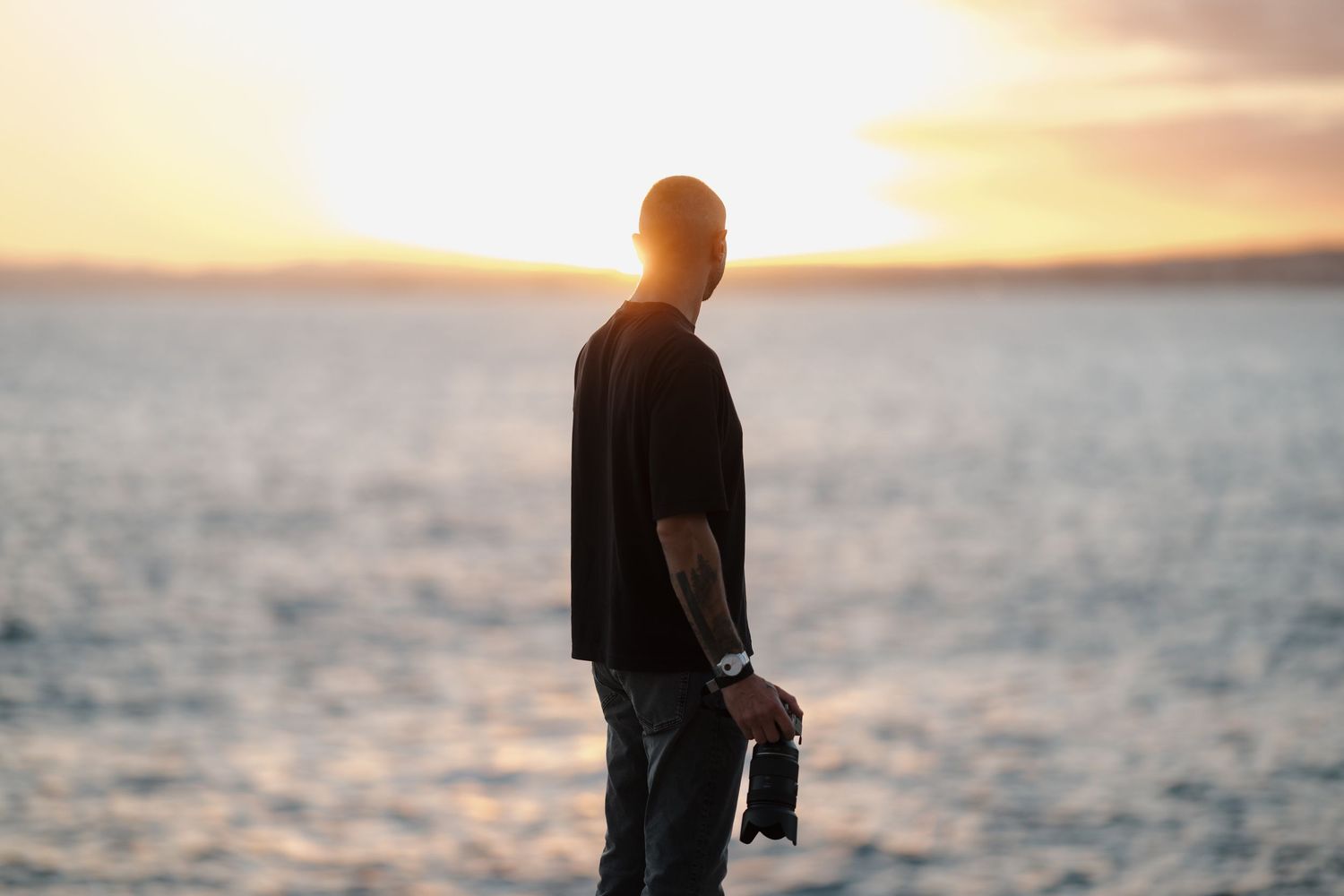 Man standing by ocean at sunset holding shoes, silhouetted against golden sky with tattoo visible on arm.