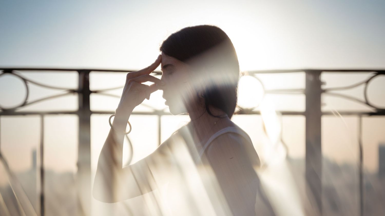 Silhouette of a woman shielding her eyes from bright sunlight on a balcony with metal railings.