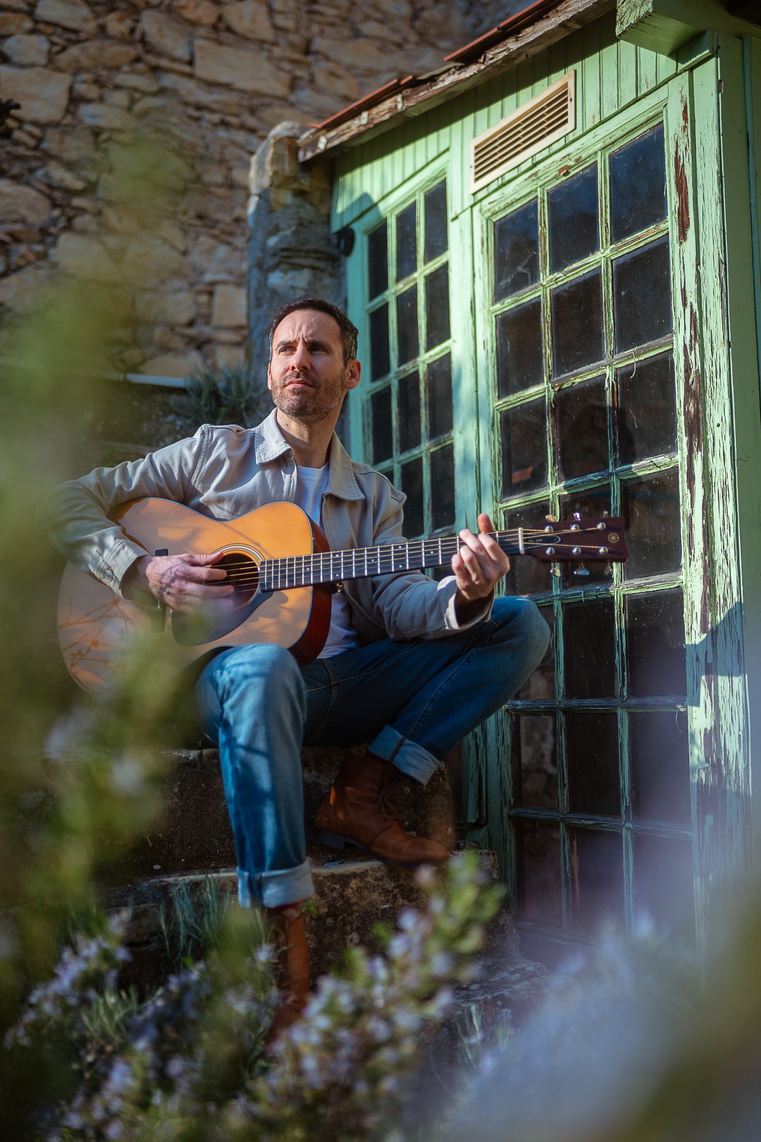 Man playing acoustic guitar seated outside rustic stone building with green vintage window.