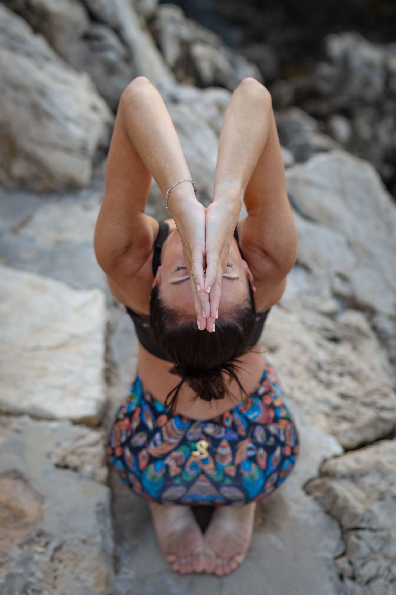 Woman in colorful skirt performing kneeling yoga pose with hands raised on rocky outdoor surface.