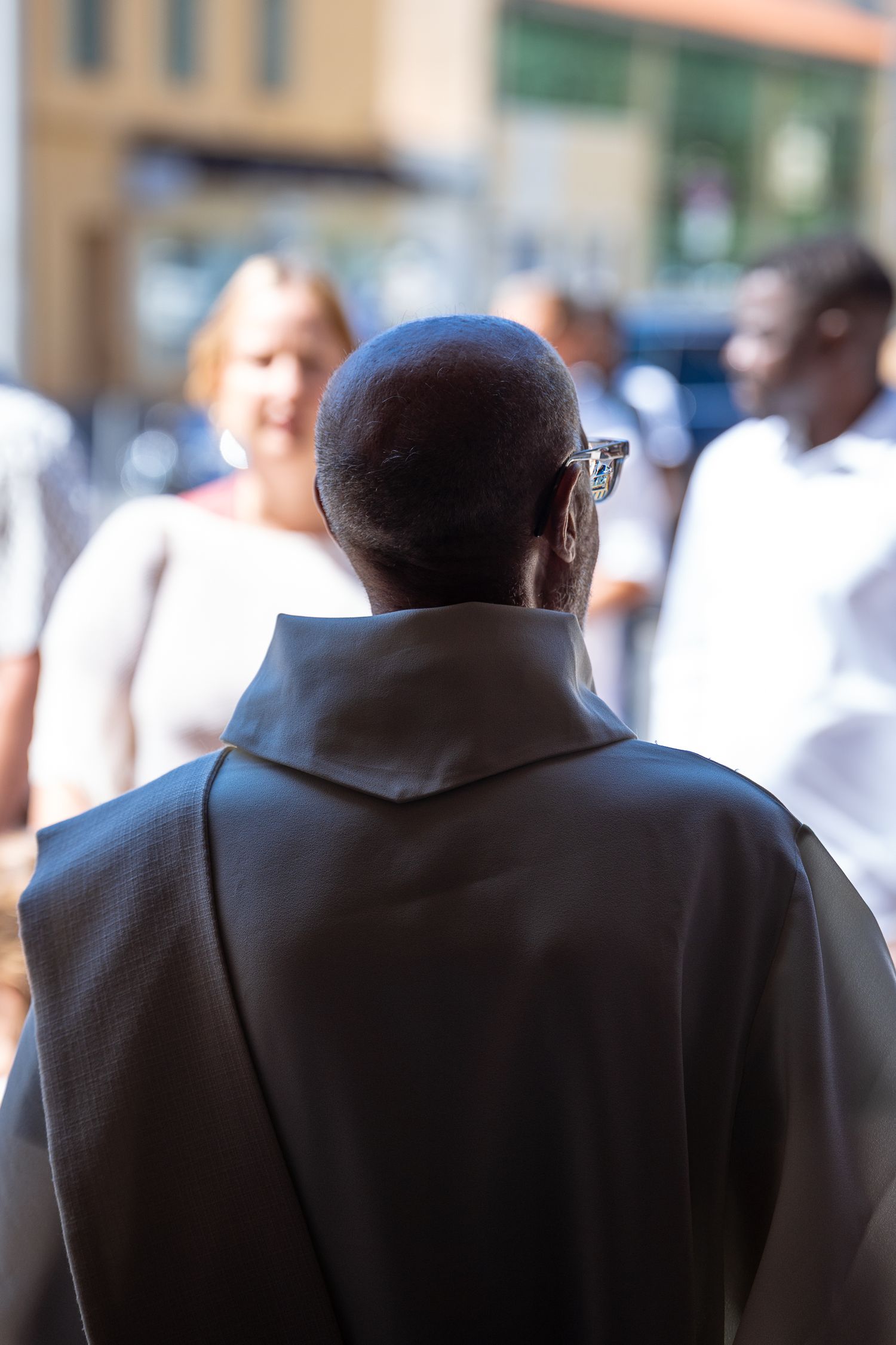 Back view of a bald priest in dark robes standing outdoors facing a group of people.