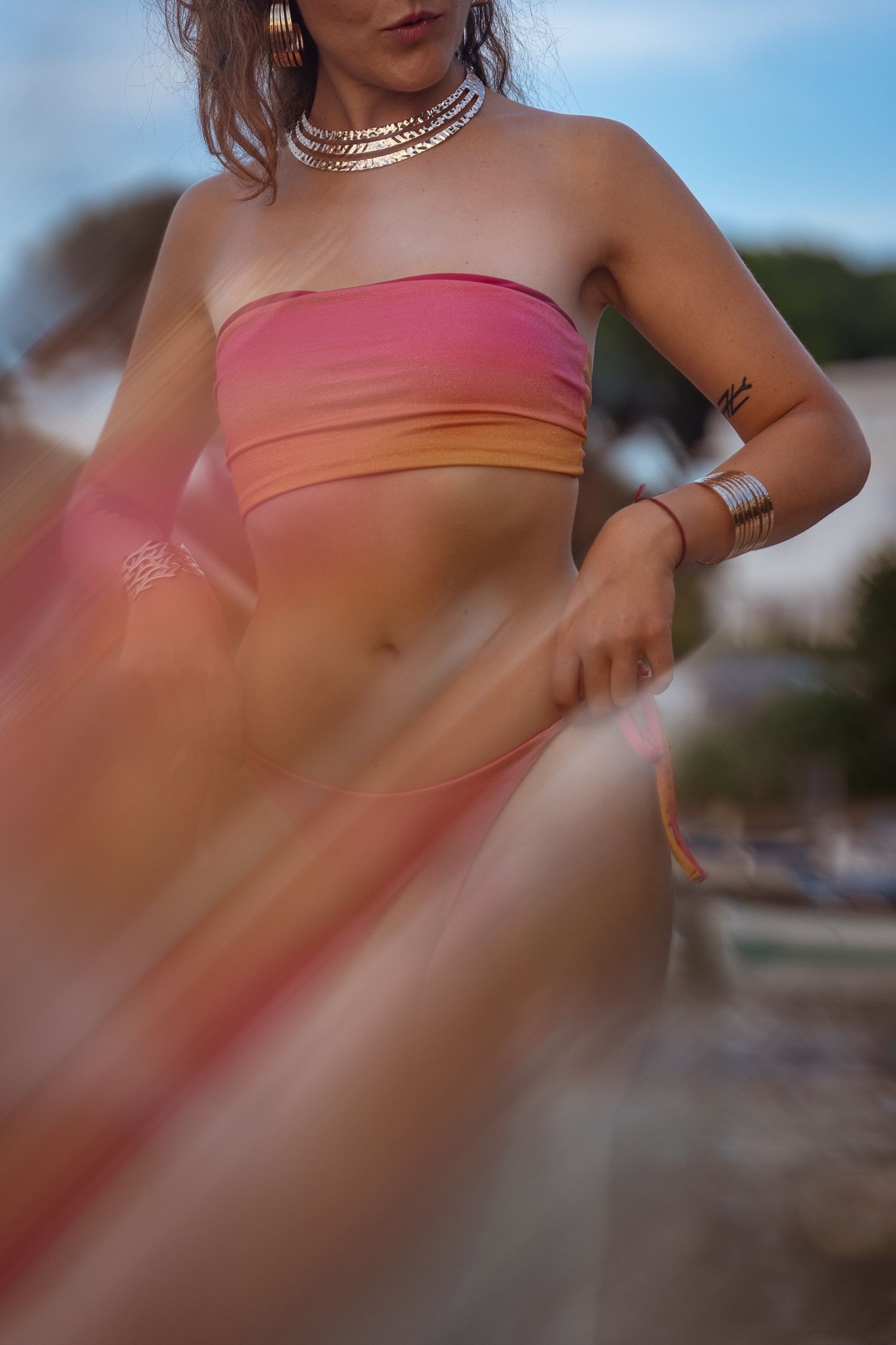 Woman in pink ombre bandeau top with silver jewelry holding flowing fabric outdoors.