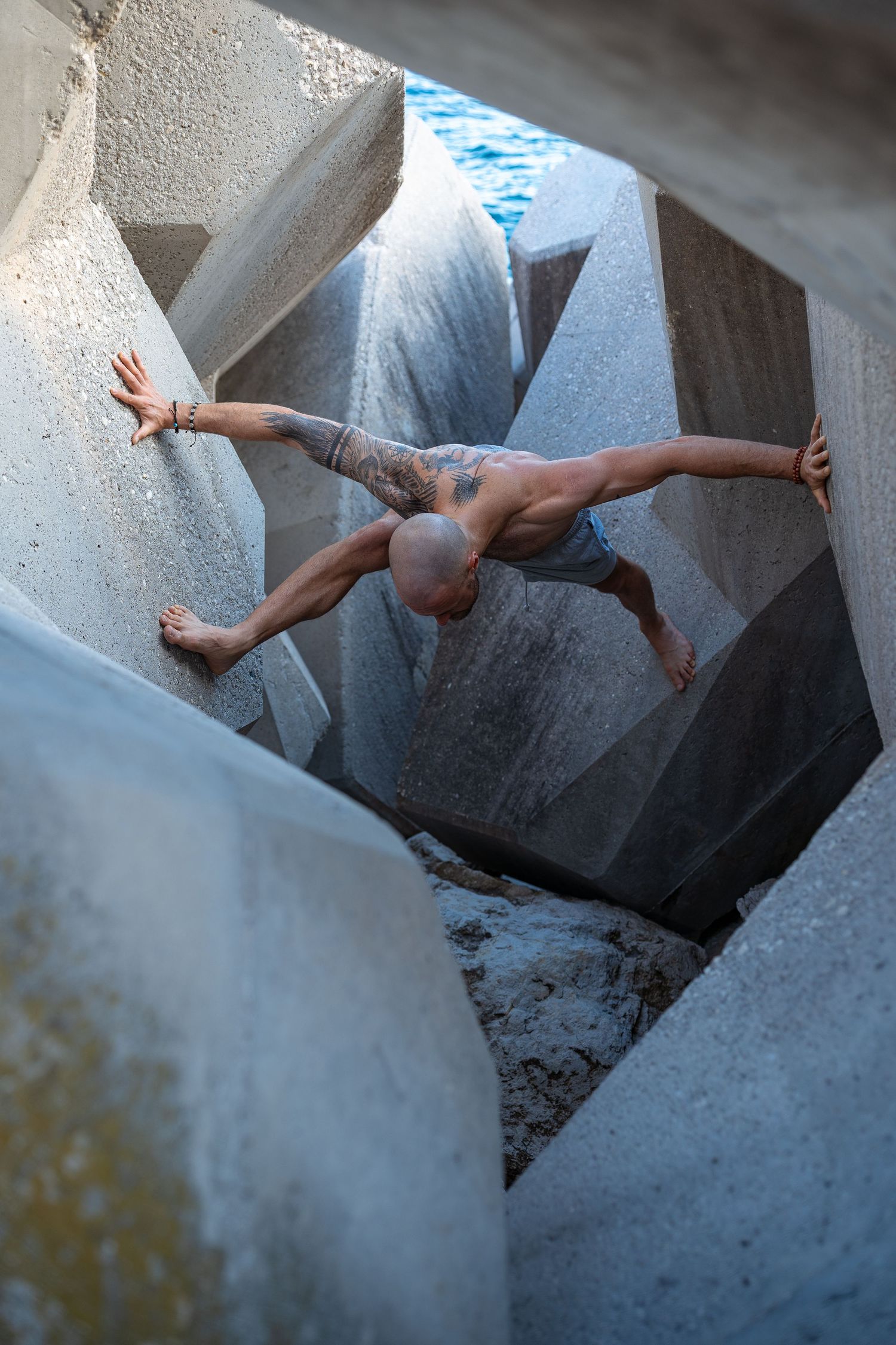Tattooed bald man balancing between large concrete tetrapod blocks near blue water.