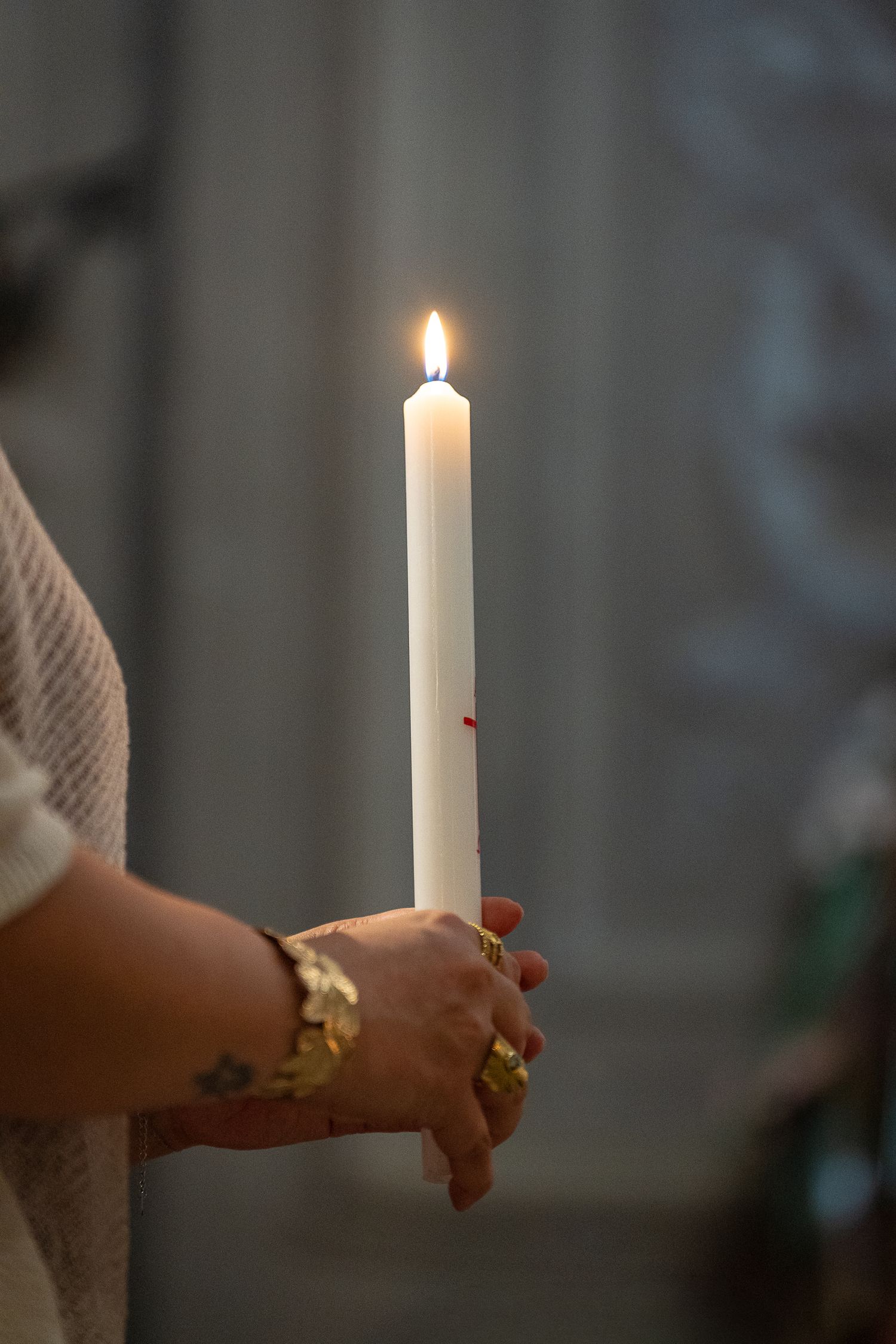 Close-up of hands holding a lit white taper candle during a religious ceremony indoors.