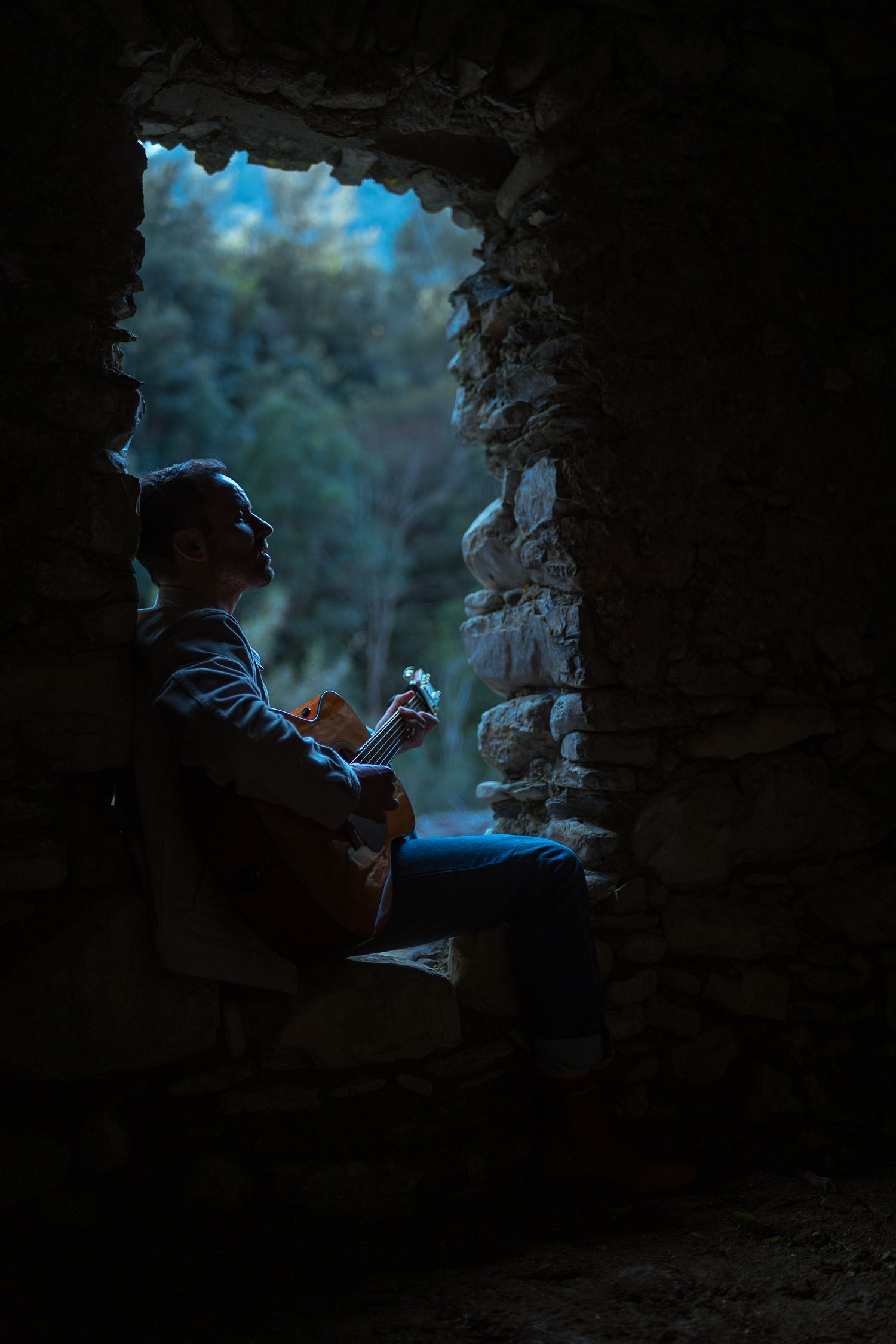 Guitarist playing acoustic guitar seated in dark stone window opening with forest view.