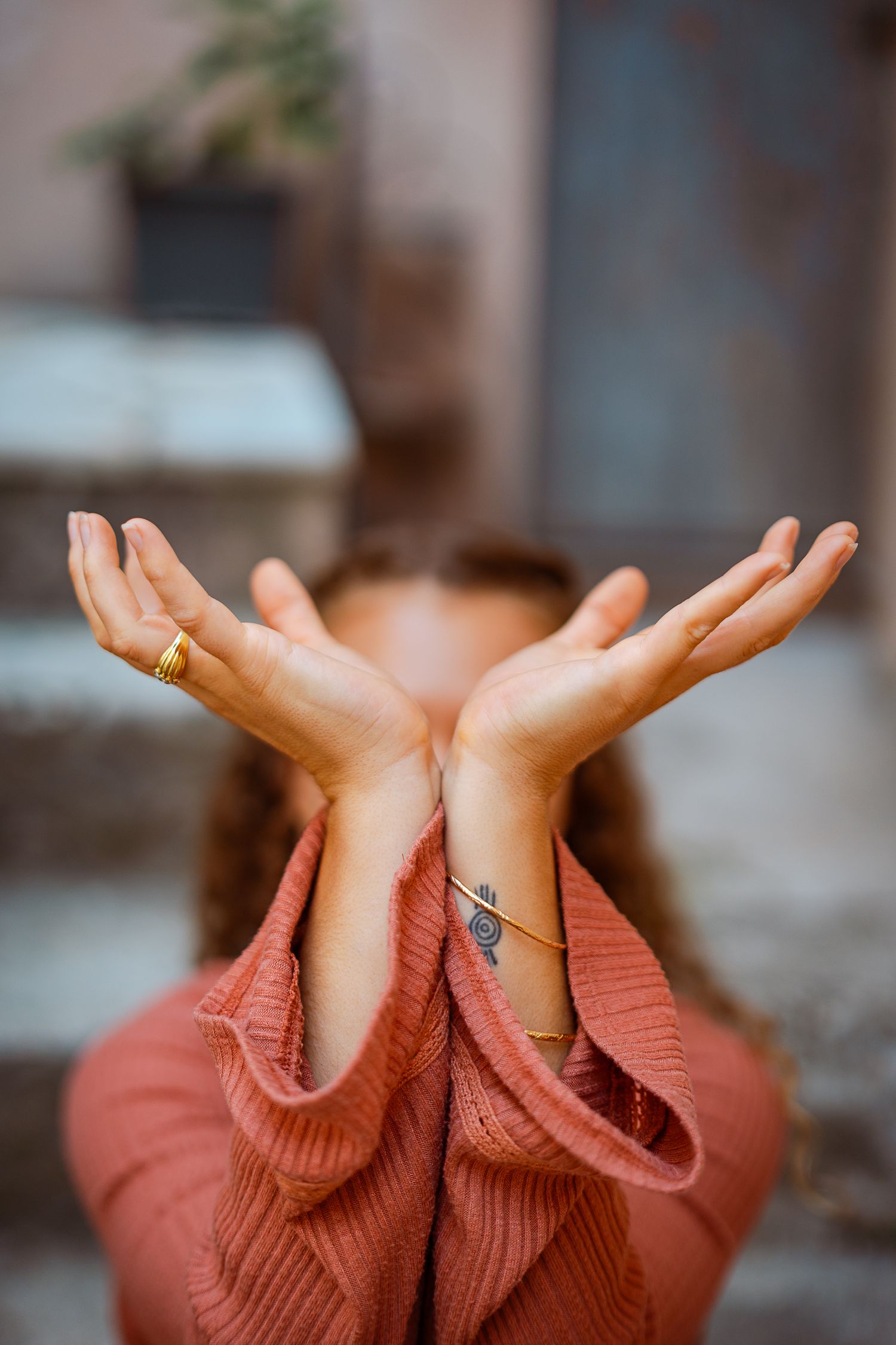 Close-up of womans crossed hands with gold rings and spiral tattoo on wrist against blurred background.
