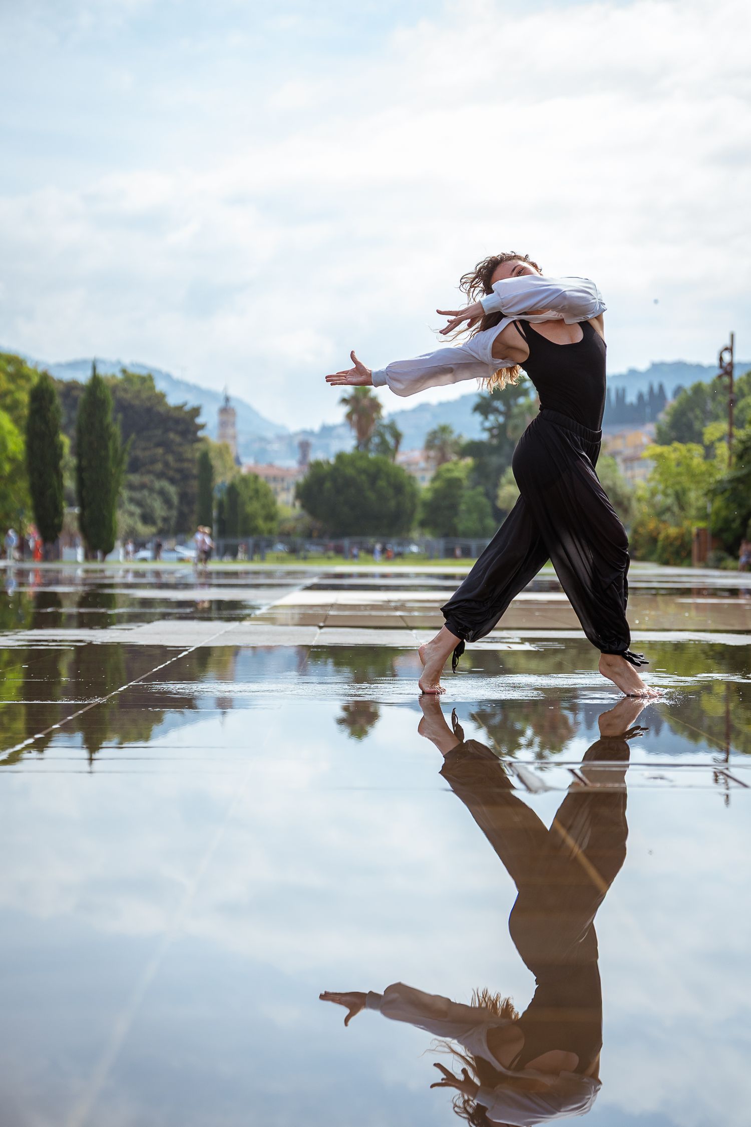 Female dancer performing expressive contemporary dance on reflective wet surface in outdoor park setting.