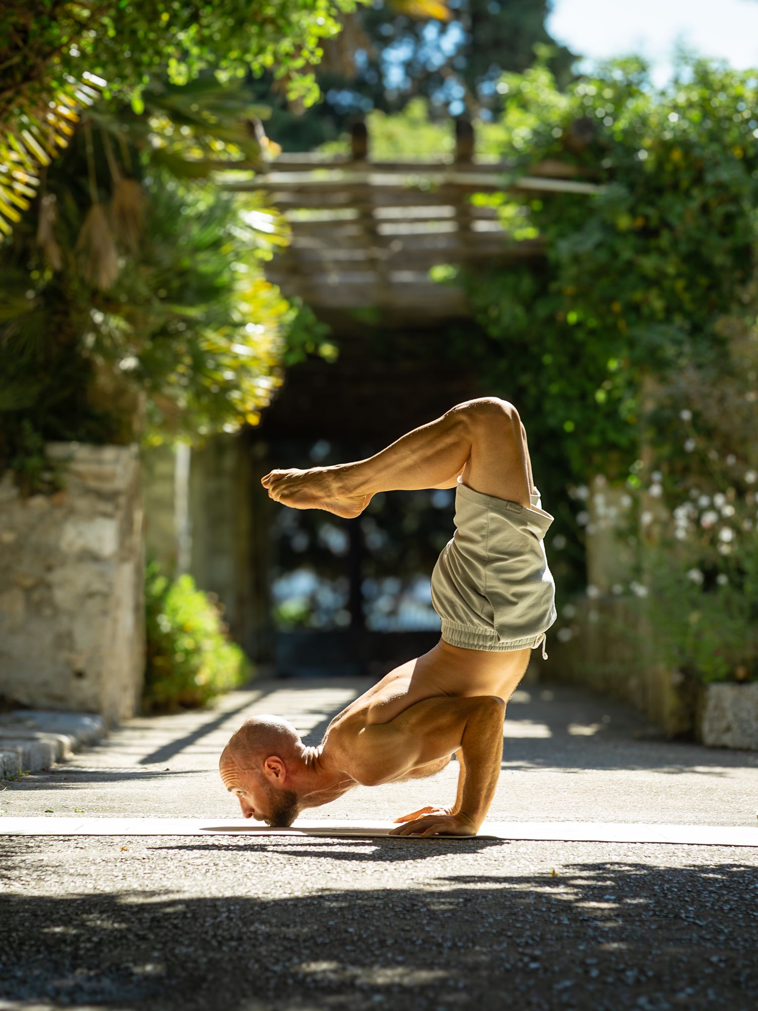 Bald shirtless man performing advanced scorpion headstand yoga pose outdoors in sunny garden setting.