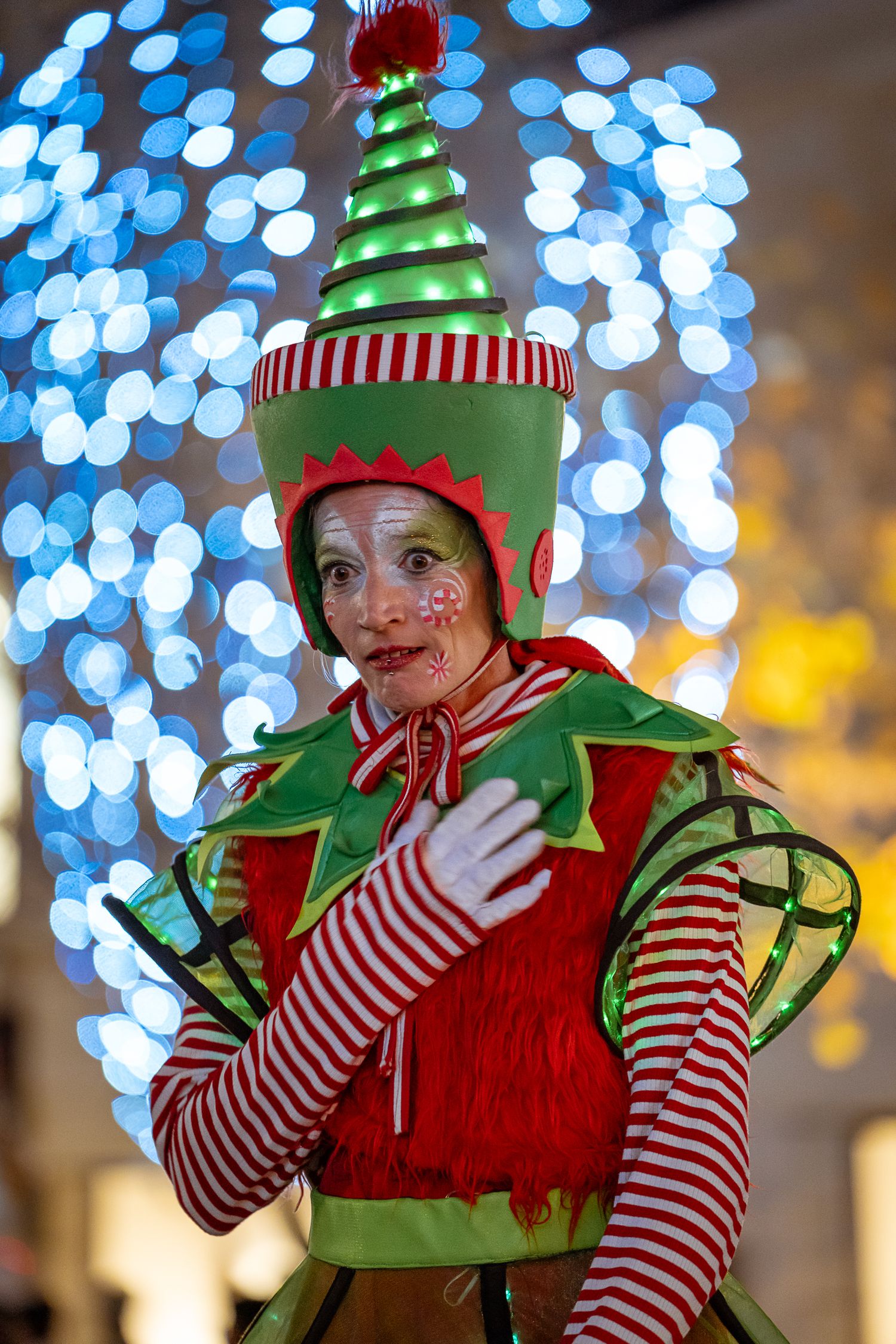 Female performer dressed in colorful Christmas elf costume with face paint posing before bokeh lights.