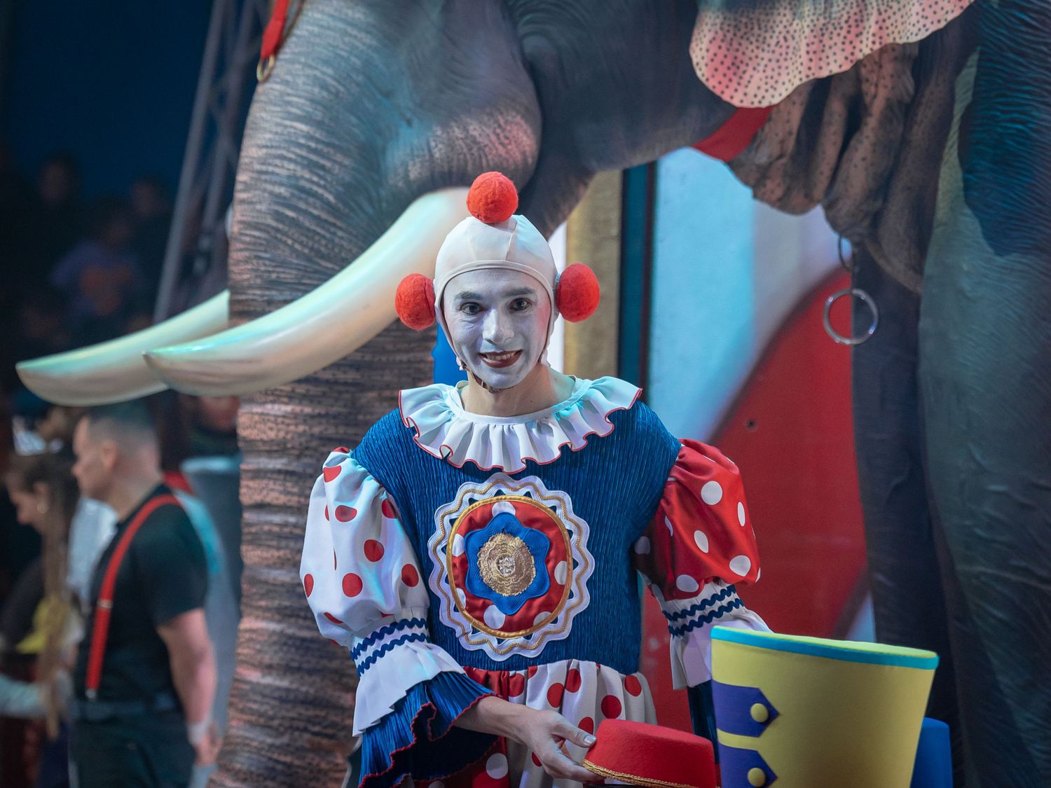 Circus clown in colorful polka dot costume standing before an elephant during a live circus performance.