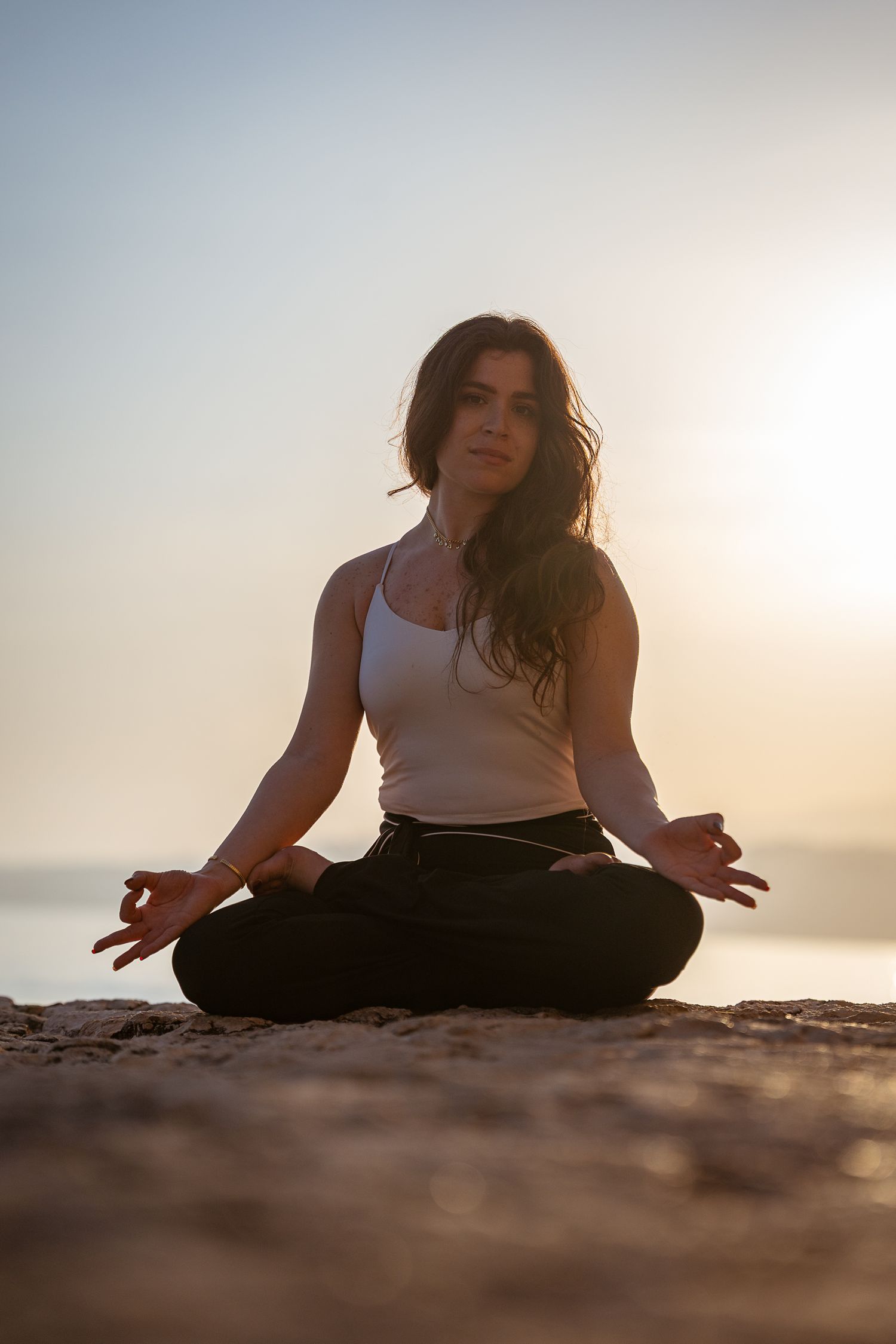 Woman in white tank top meditating in lotus pose on rocky surface during golden sunset.