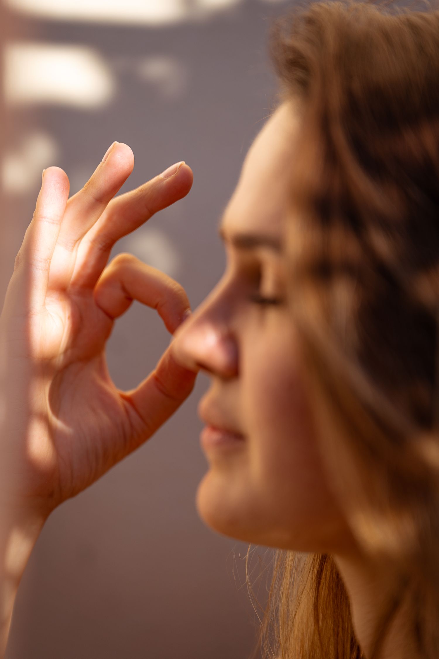 Close-up profile of young woman with eyes closed making OK gesture near nose in sunlight.