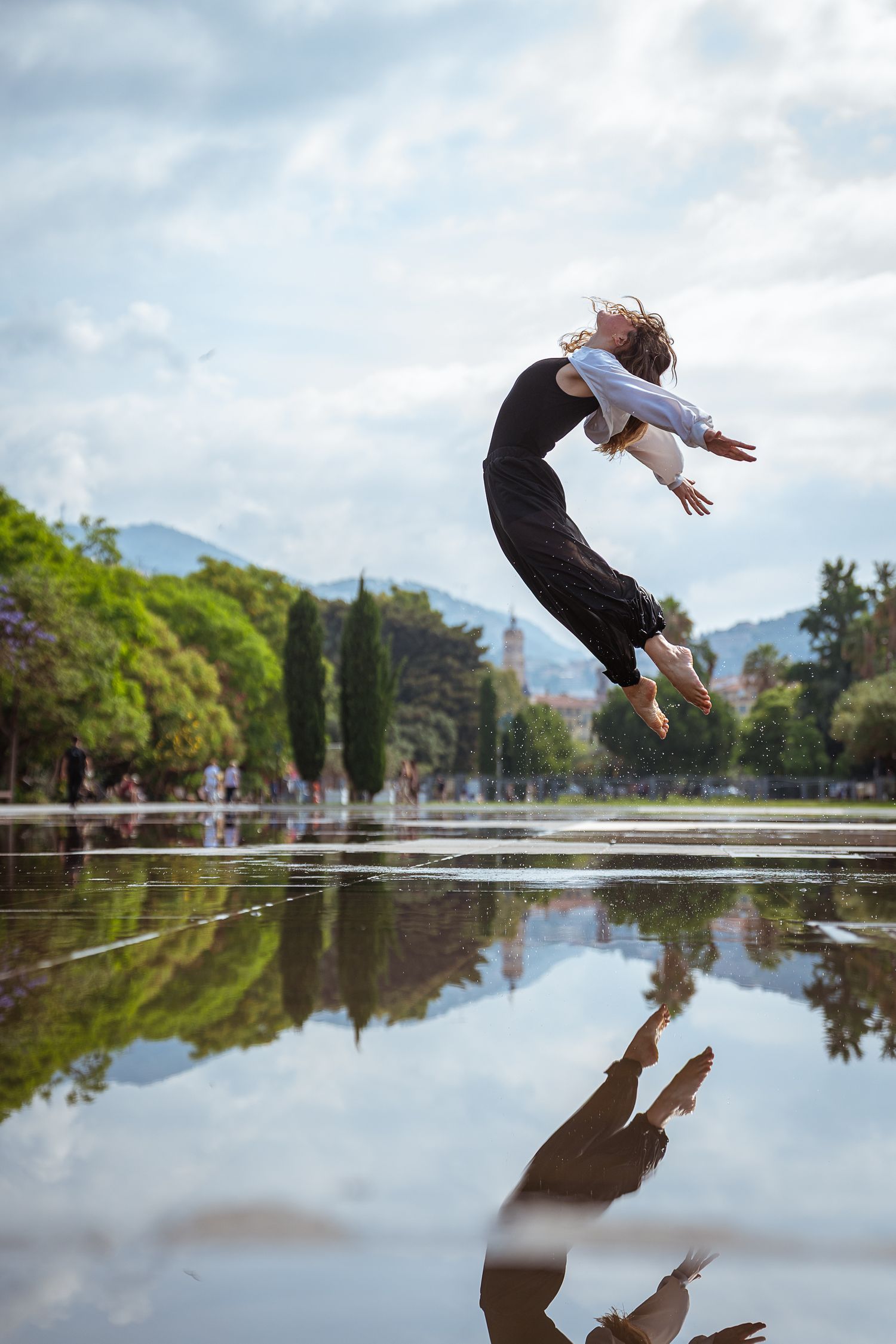 Dancer leaping mid-air over reflective wet surface in park with mountains in background.
