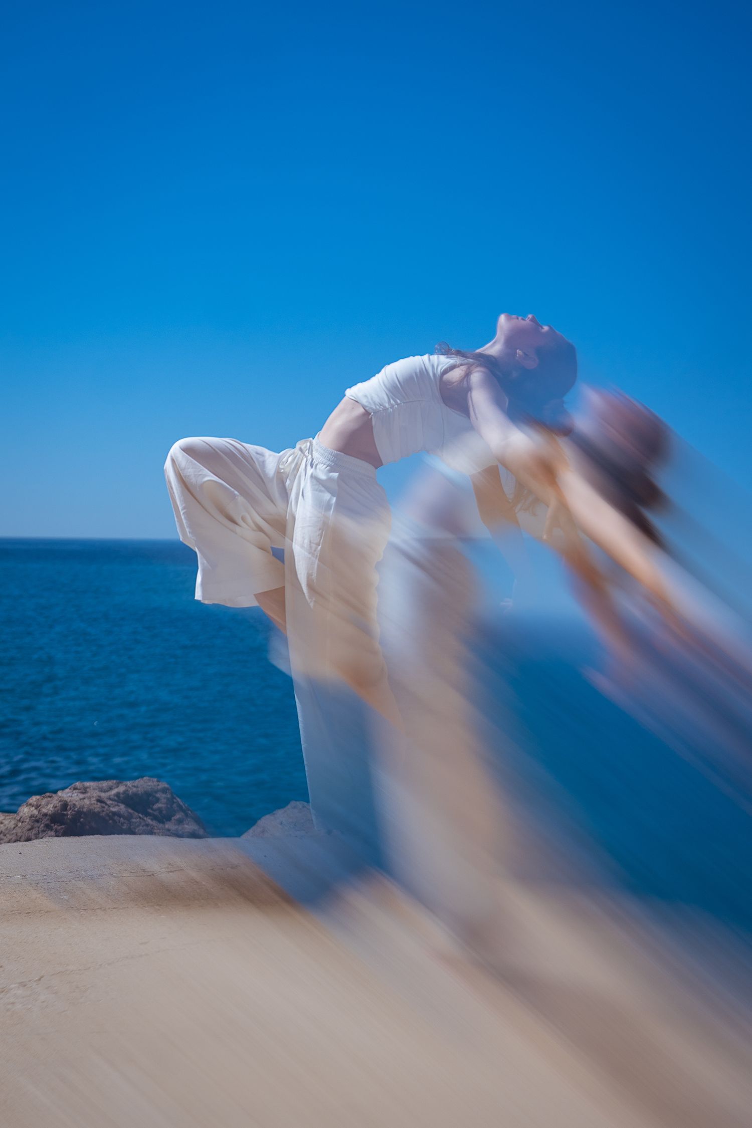 Woman in white flowing dress dancing on rocky beach with blue ocean and clear sky backdrop.