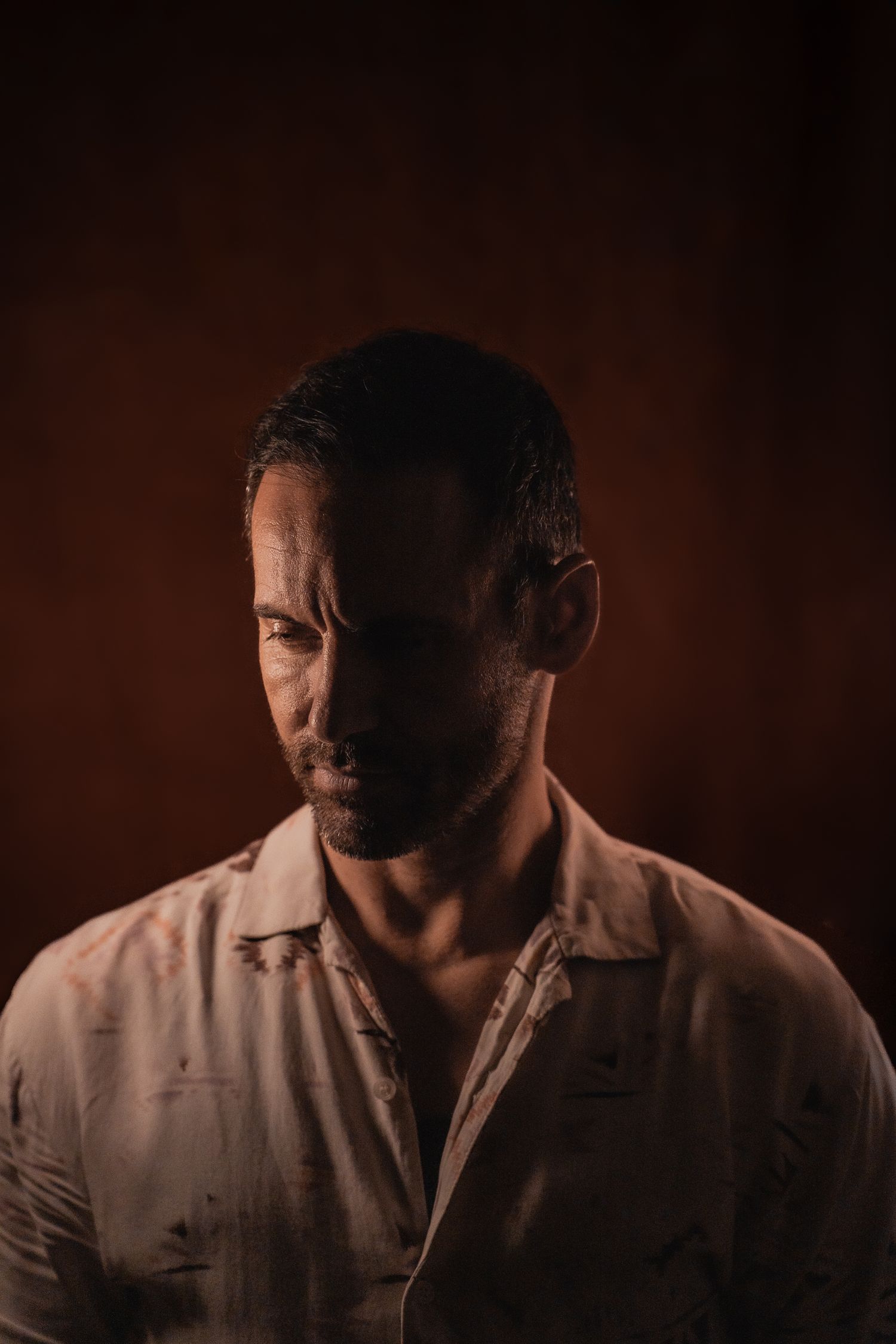 Dramatic low-light portrait of bearded man in patterned shirt against dark brown background.