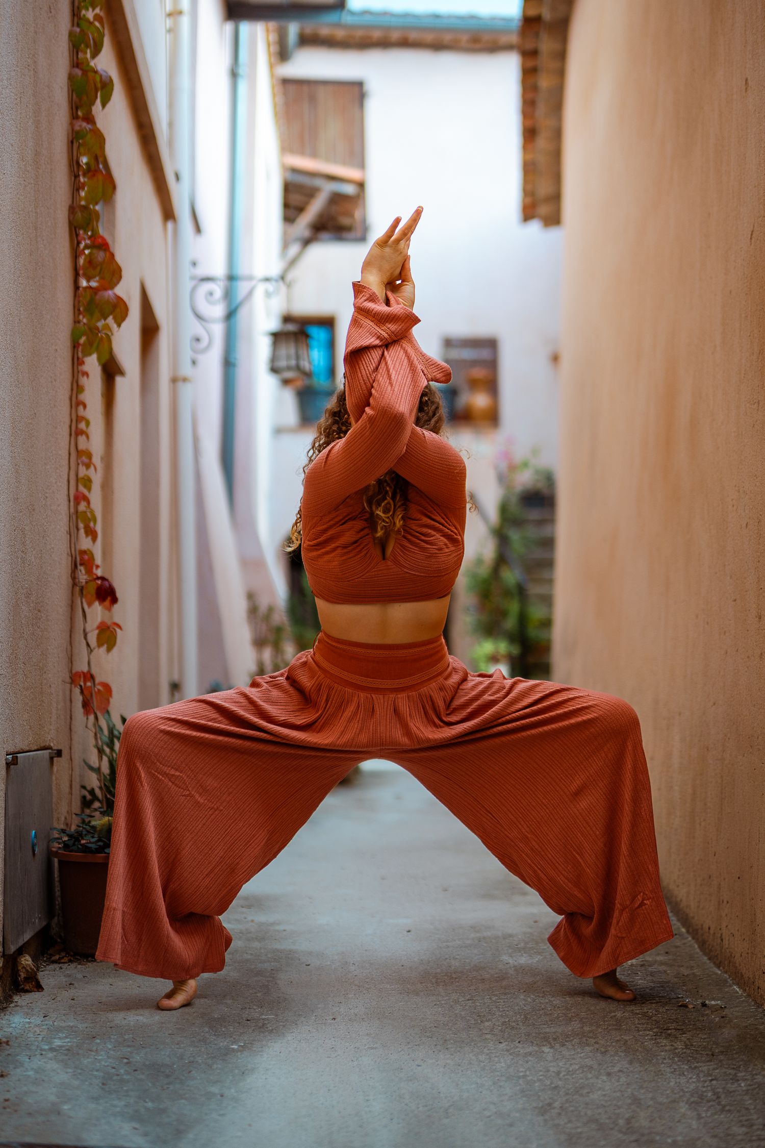 Woman in rust wide-leg pants performing goddess yoga pose in narrow European alleyway between buildings.