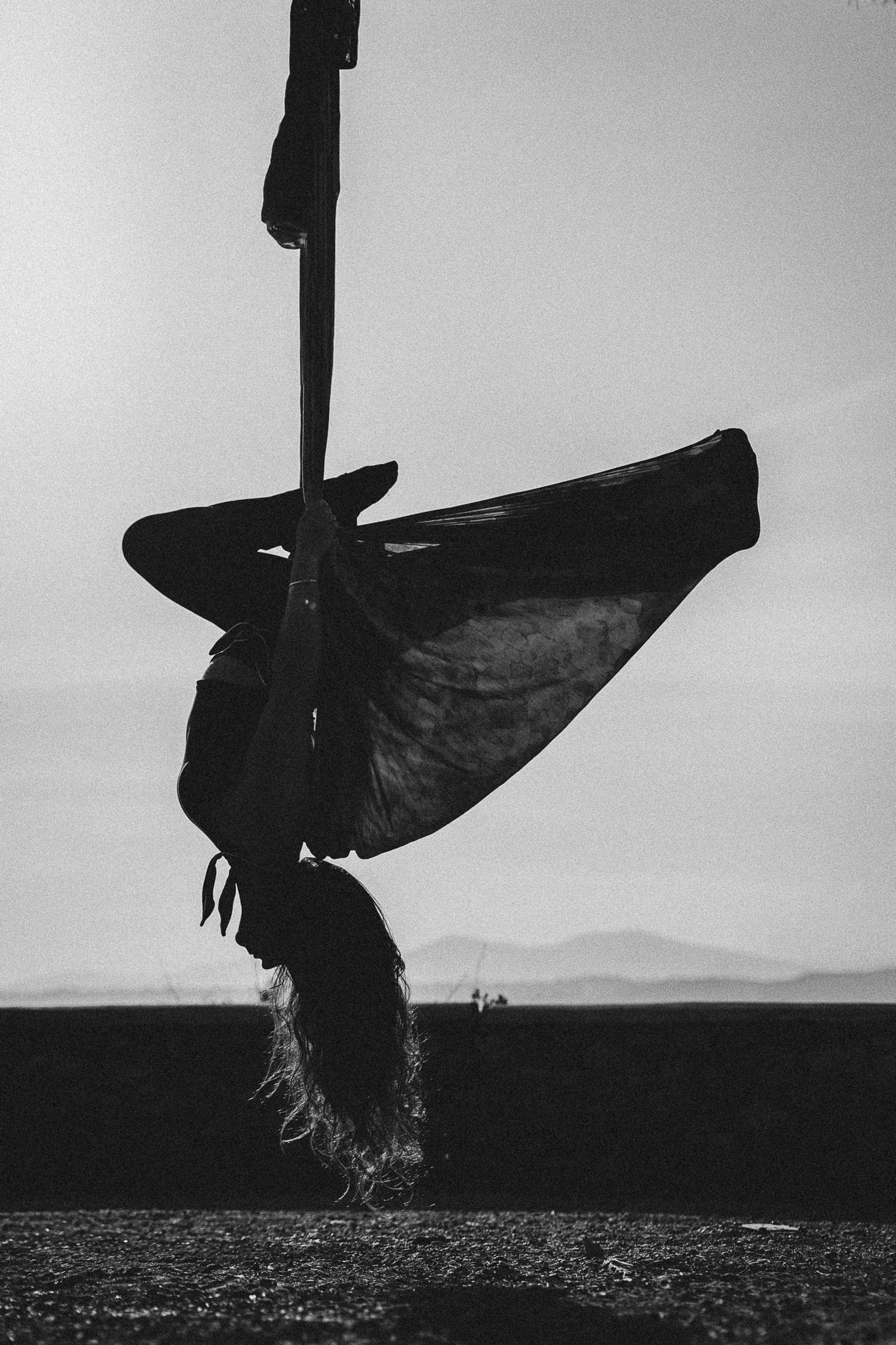 Black and white silhouette of aerial dancer hanging upside down from silk against open sky.