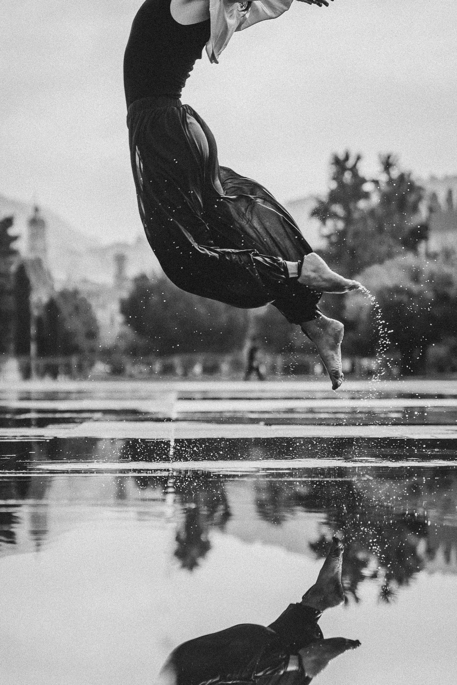 Black and white photo of dancer jumping over reflective water surface with city backdrop.