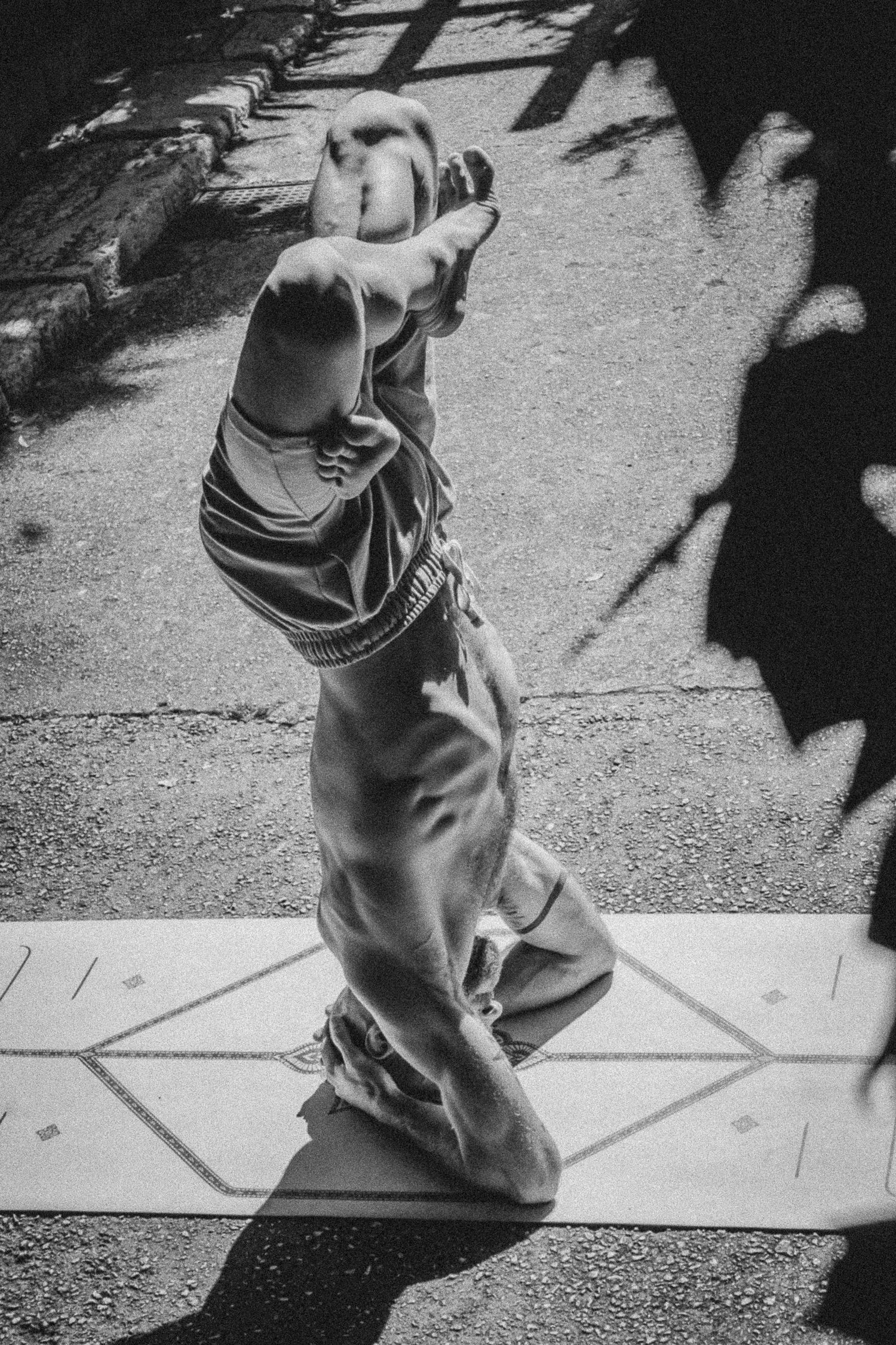 Black and white photo of person performing headstand yoga pose on mat with dramatic shadows on pavement.