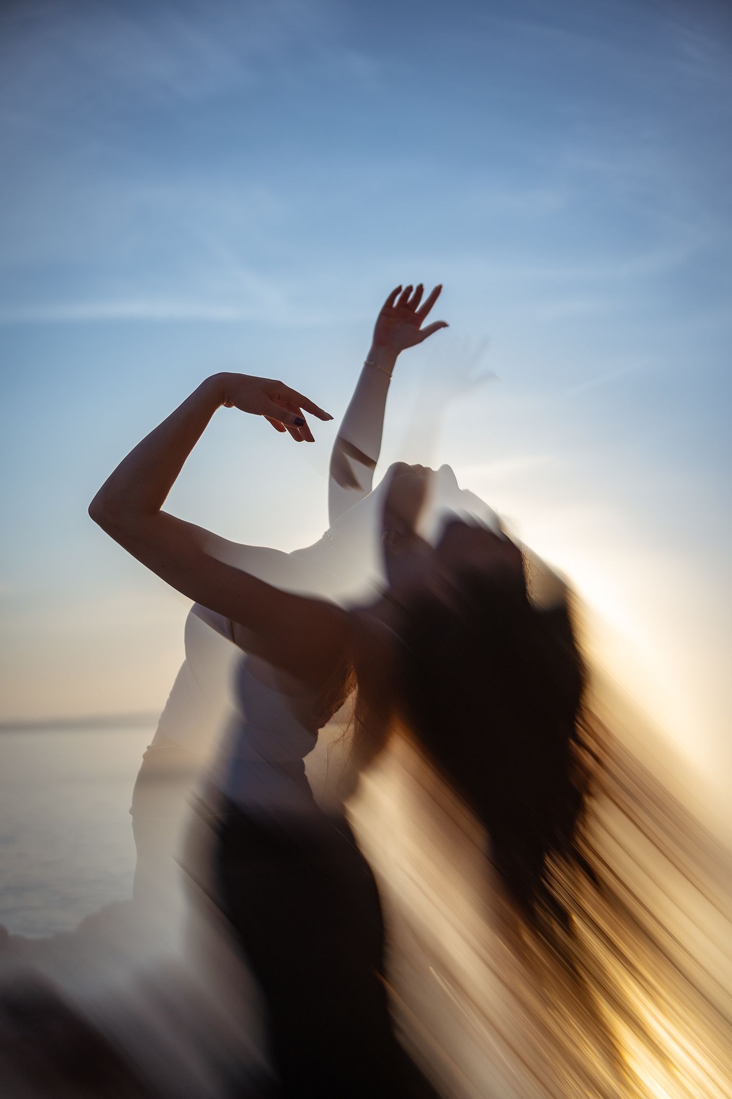 Blurred motion silhouette of woman dancing with arms raised against golden sunset sky backdrop.