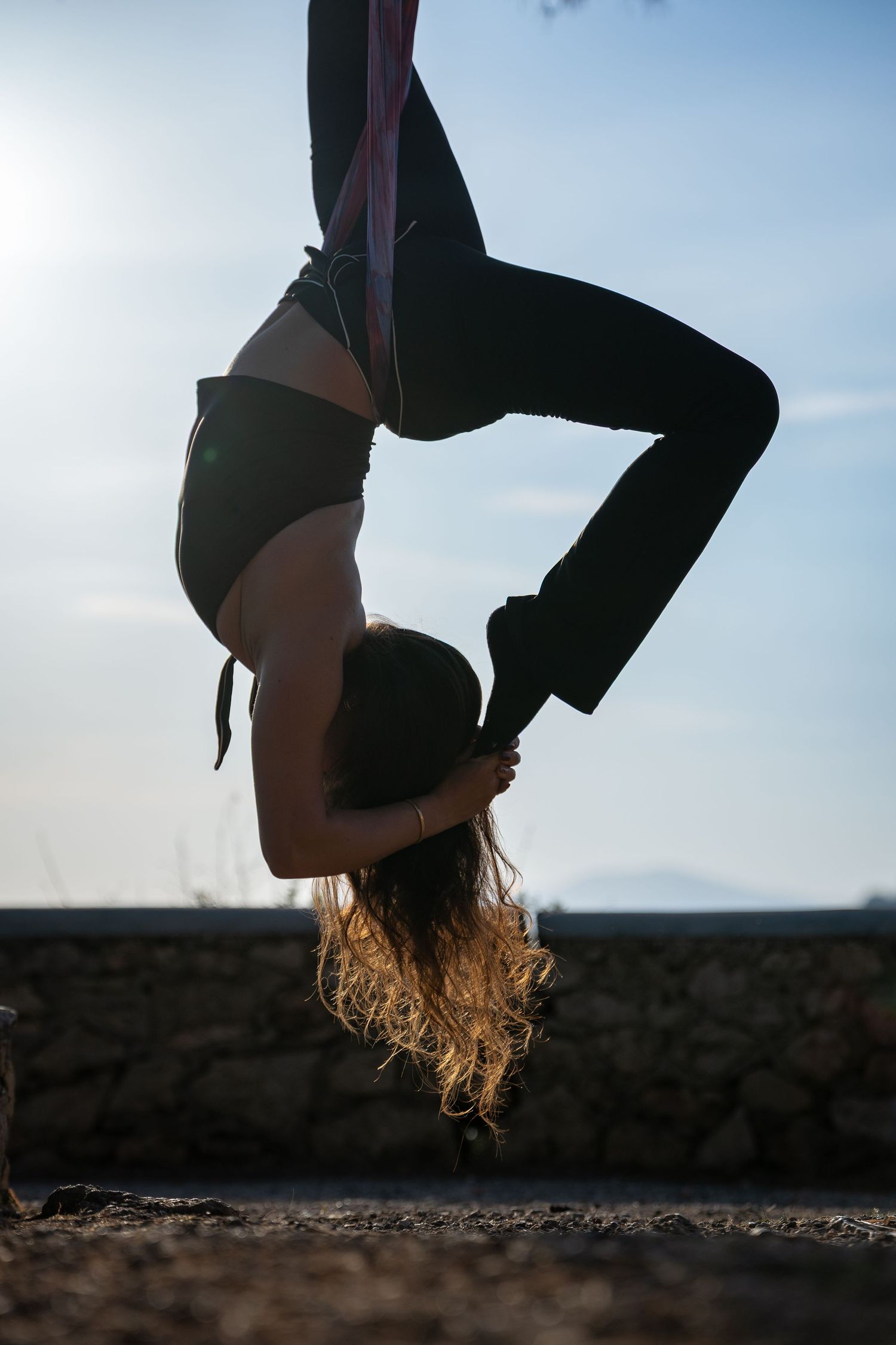 Silhouette of woman hanging upside down performing aerial yoga against bright sky.