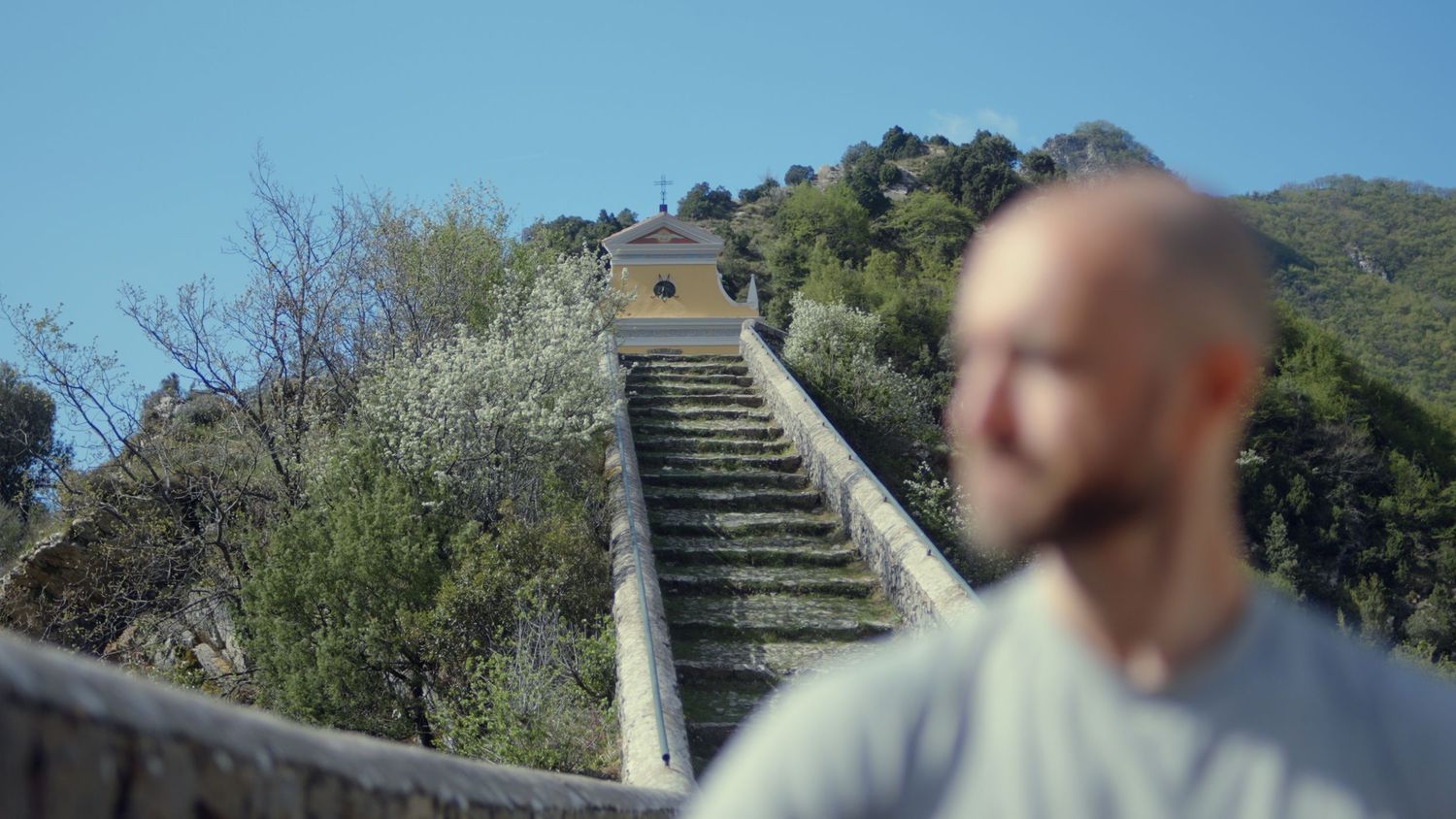 Bald man in foreground with historic stone staircase and chapel on hill behind.