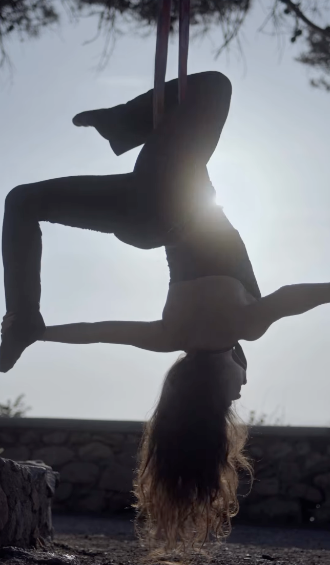 Silhouette of aerial dancer performing inverted pose on hanging silks outdoors.