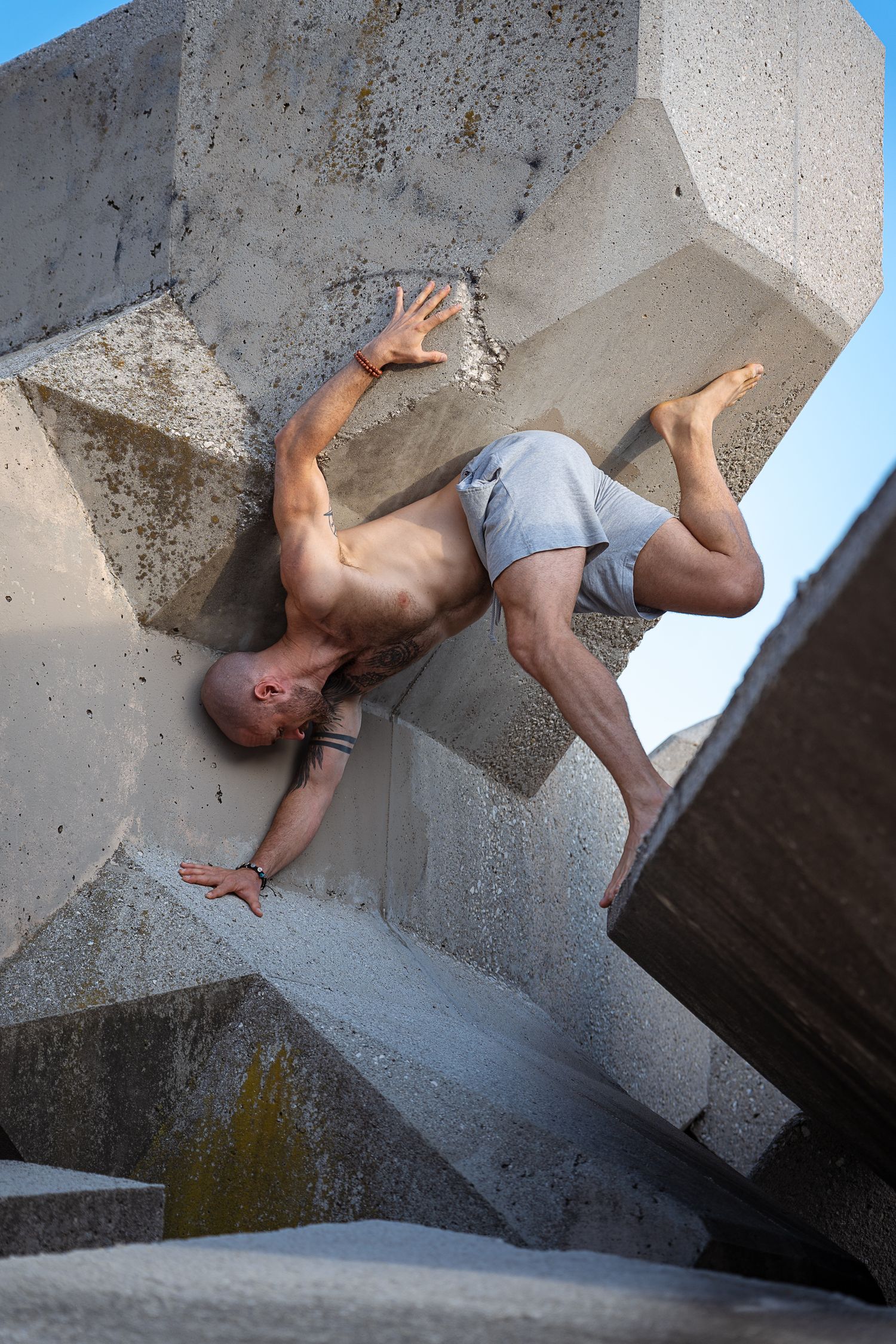 Shirtless bald man climbing and balancing on angular concrete sculpture against blue sky.